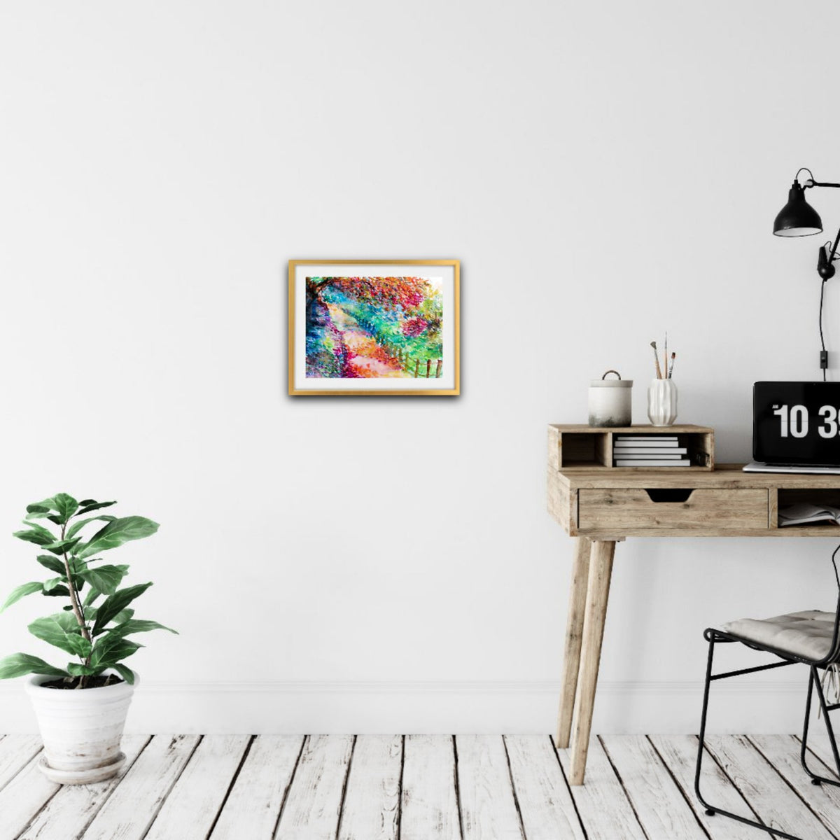 Colorful Autumn Landscape, original fall watercolor painting with an autumn path on paper by contemporary artist TRAXI. A colorful abstract framed wall art print hangs on a white wall in a modern home office setting with a wooden desk, black lamp, and a potted green plant on a light wood floor.