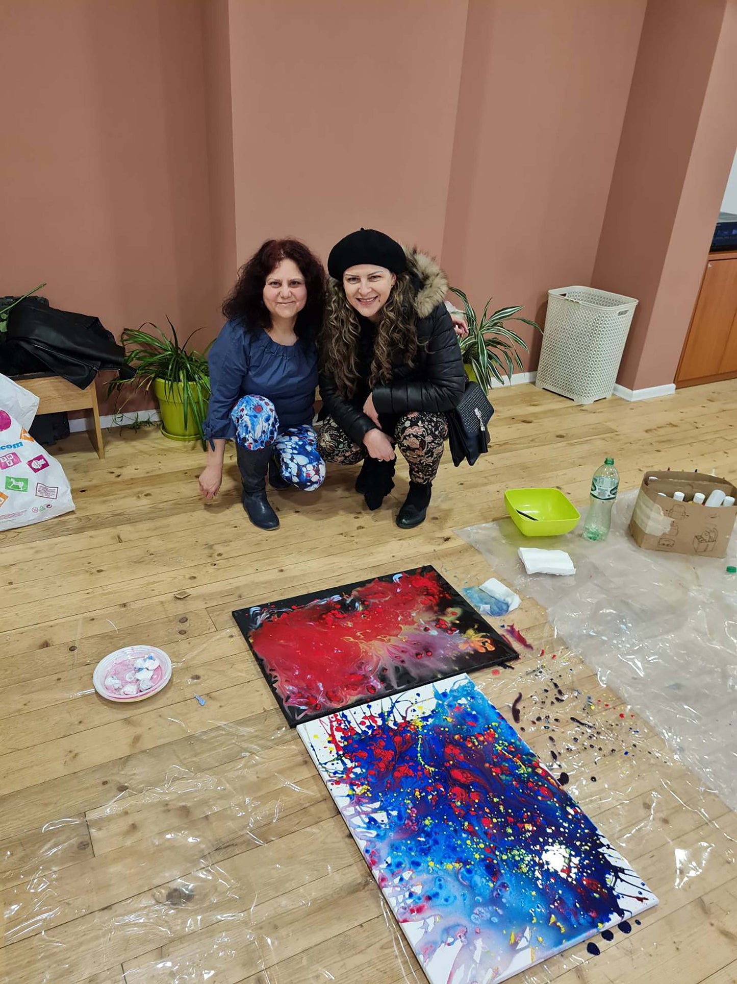 Mood of the Party original vertical abstract fluid acrylic painting with a vibrant blue wave and splashes of red and yellow by Diana Dimova TRAXI ART. Two women crouching on a wooden floor in a studio, smiling at the camera with two colorful abstract paint splatter canvases in front of them. The room has dusty pink walls, and various art supplies like paint palettes, brushes, and plastic sheeting are scattered around.