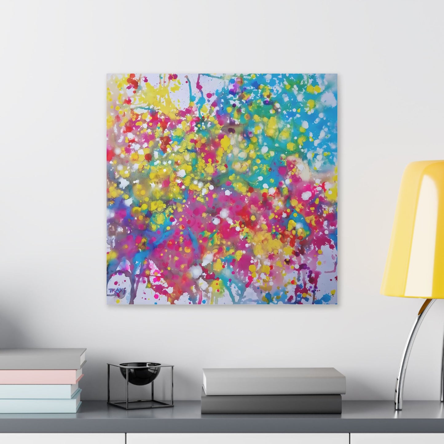 Painting of happiness 2 vibrant abstract canvas print by award-winning contemporary artist Diana Dimova - TRAXI, featuring radiant pink, yellow, and blue colors. A square abstract canvas print with a vibrant splatter pattern in pink, yellow, blue, green, and purple, displayed on a white wall above a gray console table with books and a yellow lamp.