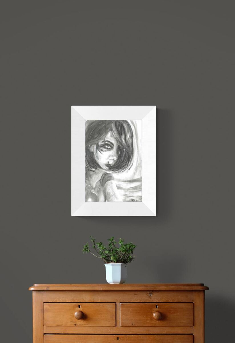 A framed black and white sketch of a girl's face hanging on a dark gray wall above a wooden dresser with a small potted plant on top. The Gaze Says It All original monochromatic charcoal portrait of a woman looking directly forward with a fearless gaze by Diana Dimova TRAXI ART.