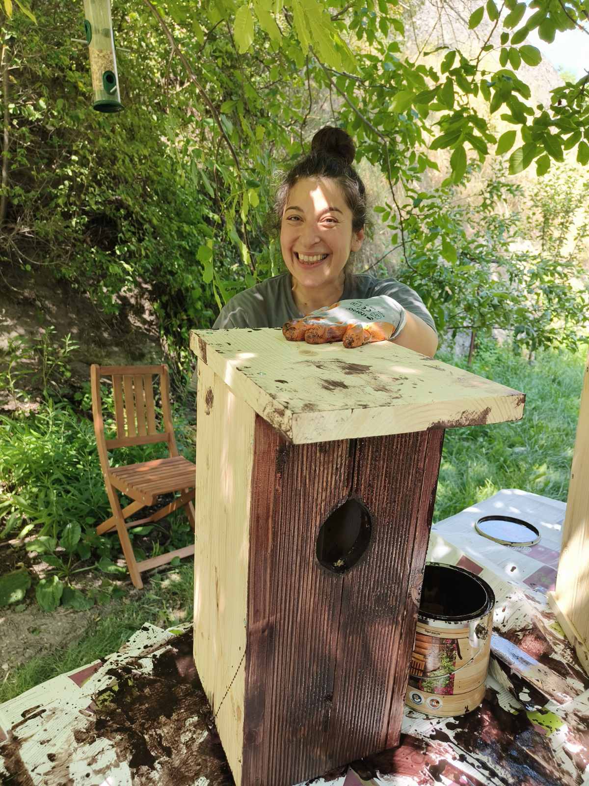 ART CAN BECOME ACTION Art Project Turning Point by Diana Dimova TRAXI A woman smiling outdoors while painting a wooden birdhouse. The birdhouse has a dark brown stained front panel, a light natural wood top, and a black oval entrance hole.