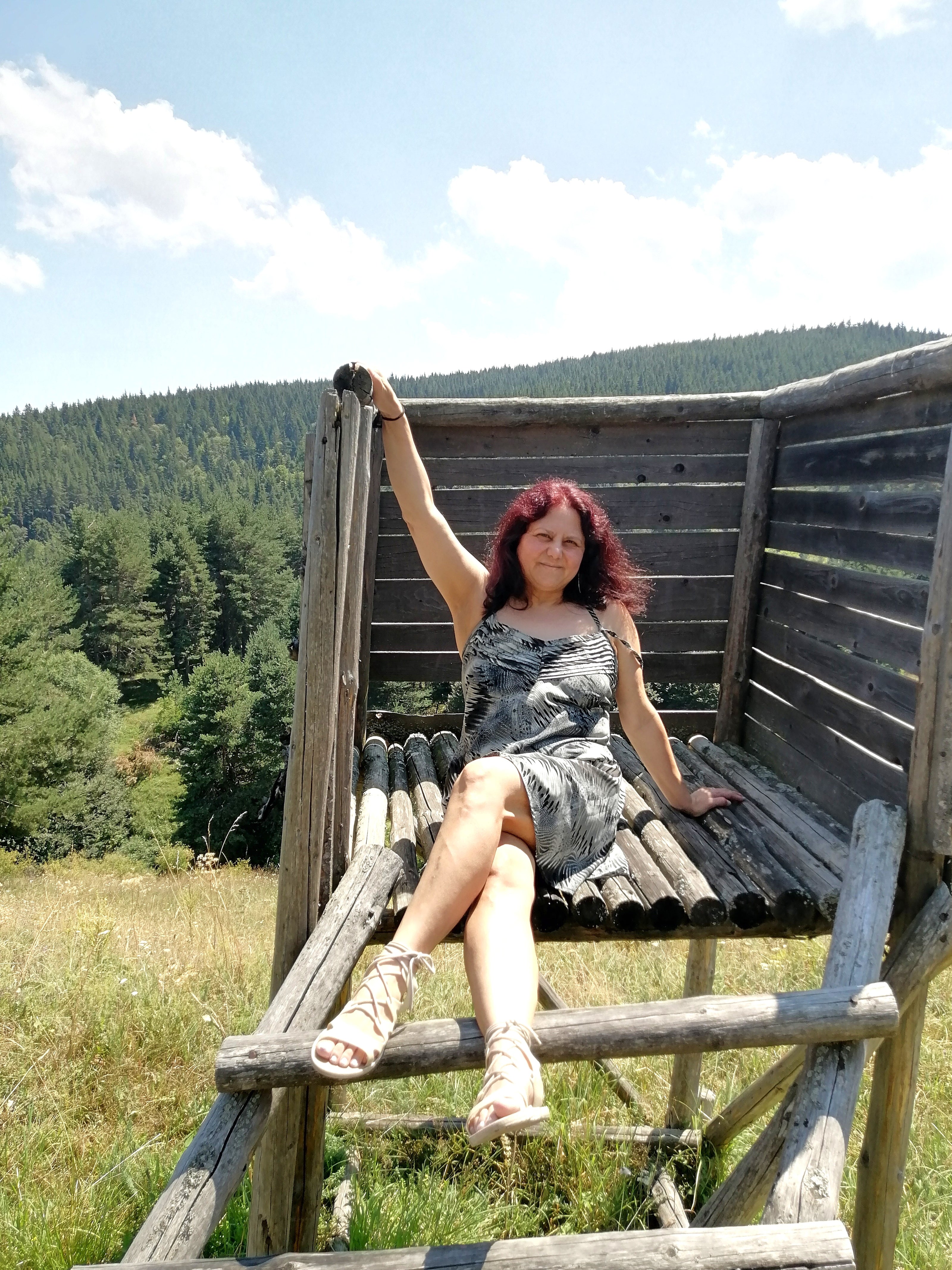 LIFE IS WONDERFUL — A Virtual Fine Art Solo Exhibition by Diana Dimova TRAXI. Art Project inspired by Nature, exploring Time. A woman with red hair wearing a gray patterned dress and beige sandals sits on a rustic wooden elevated bench in an outdoor mountain landscape with trees and blue sky.