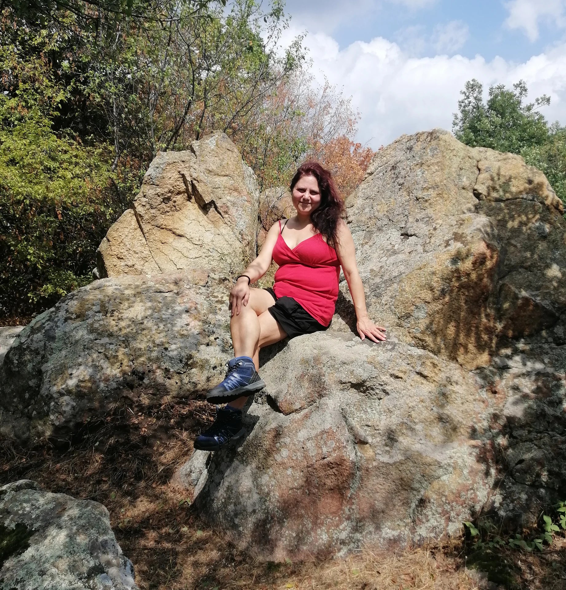 LIFE IS WONDERFUL — A Virtual Fine Art Solo Exhibition by Diana Dimova TRAXI. Art Project inspired by Nature, exploring Time. A woman wearing a red tank top and black shorts sits on a large rock in an outdoor forest setting with trees and sky visible.