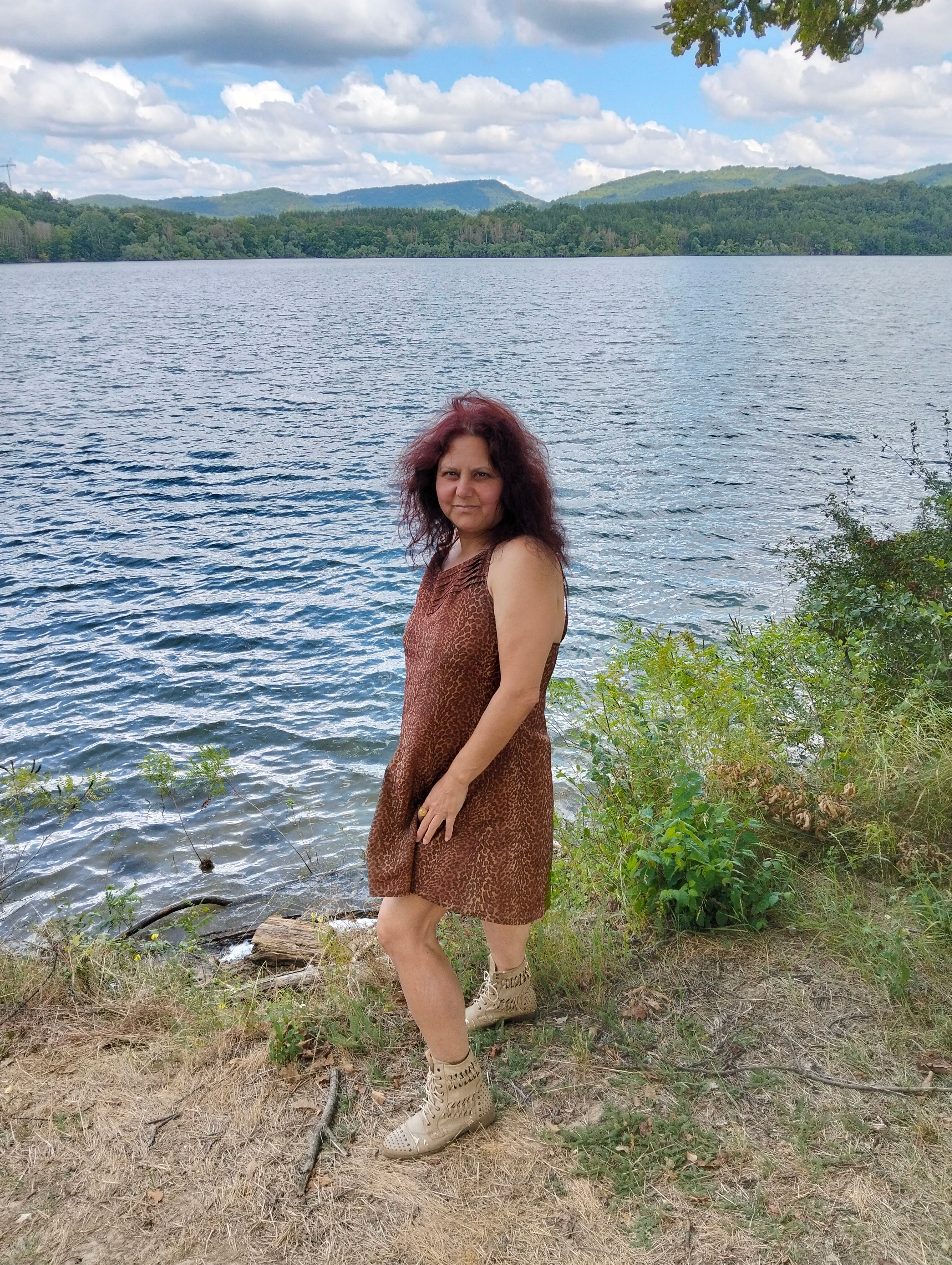 LIFE IS WONDERFUL — A Virtual Fine Art Solo Exhibition by Diana Dimova TRAXI. Art Project inspired by Nature, exploring Time.

 A woman with reddish hair stands on a grassy lakeshore wearing a sleeveless brown patterned dress and beige boots, with a large lake, distant green hills, and a cloudy blue sky behind her.