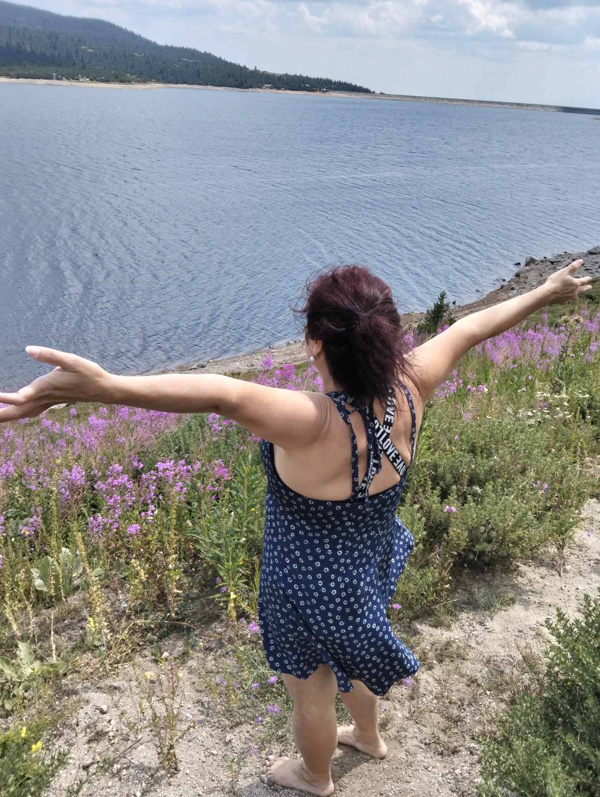 LIFE IS WONDERFUL — A Virtual Fine Art Solo Exhibition by Diana Dimova TRAXI. Art Project inspired by Nature, exploring Time. A woman stands barefoot outdoors by a large body of water, facing away from the camera with her arms outstretched. She is wearing a dark navy sleeveless dress with a small white floral pattern and black-and-white striped shoulder straps.