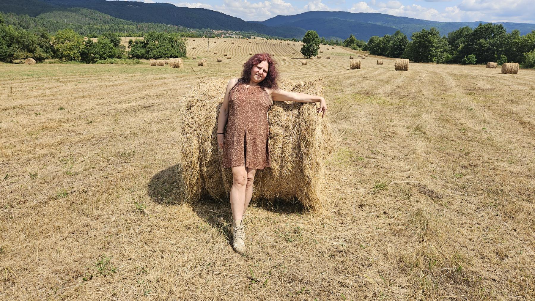 LIFE IS WONDERFUL — A Virtual Fine Art Solo Exhibition by Diana Dimova TRAXI. Art Project inspired by Nature, exploring Time.
A woman with red hair wearing a brown knit dress stands next to a large hay bale in a dry, harvested field with mountains in the background.