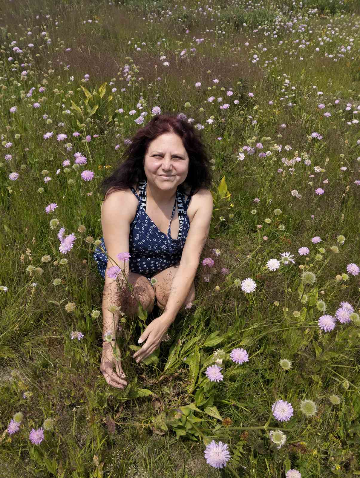 LIFE IS WONDERFUL — A Virtual Fine Art Solo Exhibition by Diana Dimova TRAXI. Art Project inspired by Nature, exploring Time. Artist TRAXI A woman with dark hair kneeling in a grassy field filled with small pink and white wildflowers, wearing a sleeveless navy top with a white pattern.