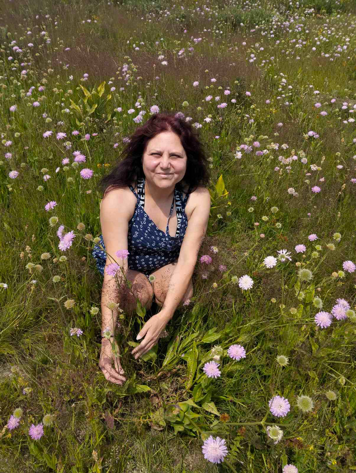 LIFE IS WONDERFUL — A Virtual Fine Art Solo Exhibition by Diana Dimova TRAXI. Art Project inspired by Nature, exploring Time. Artist TRAXI A woman with dark hair kneeling in a grassy field filled with small pink and white wildflowers, wearing a sleeveless navy top with a white pattern.