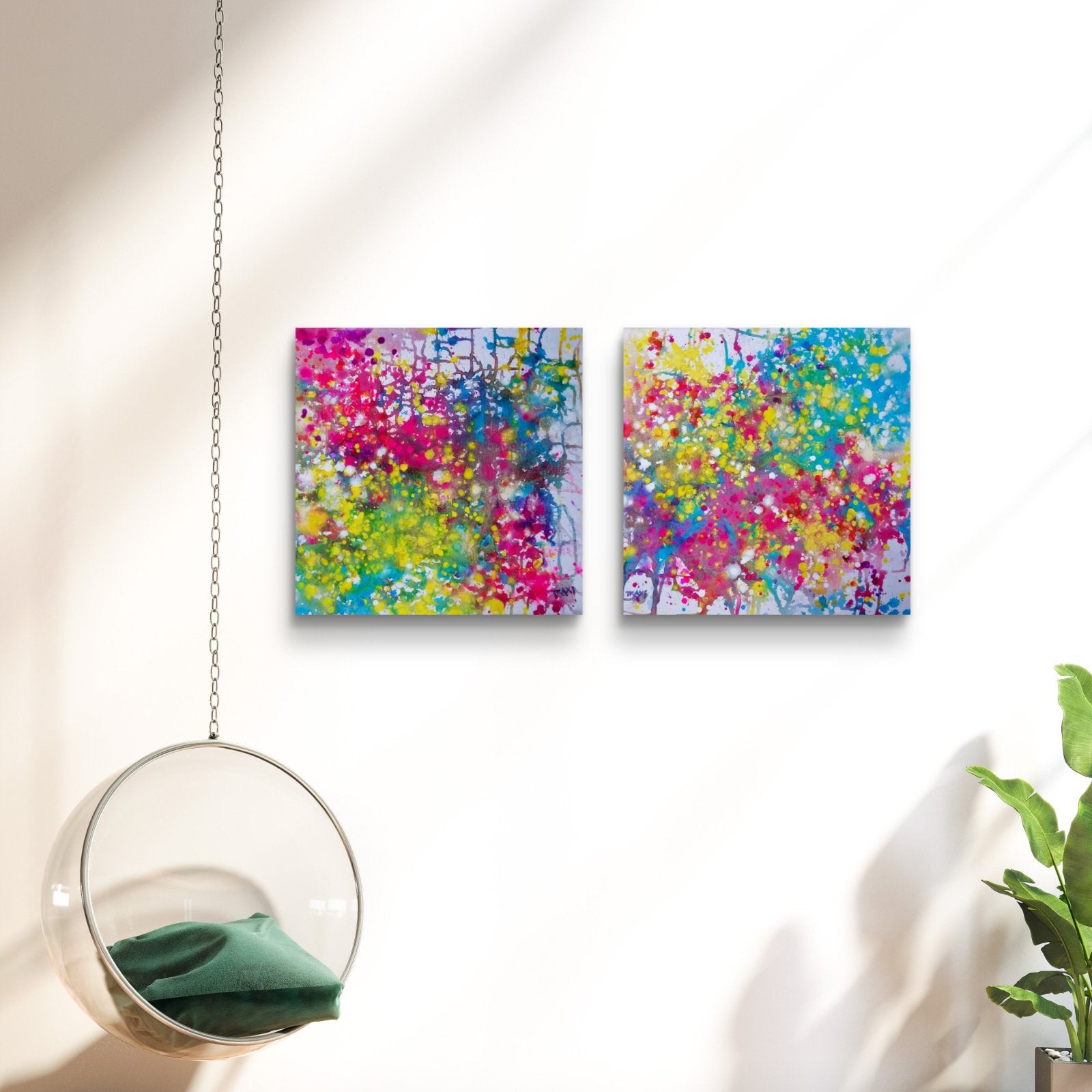 Paintings of Happiness Dyptich by Diana Dimova TRAXI,  Colorful Abstract Art Set. Two square abstract wall art prints with vibrant splashes of pink, yellow, blue, green, and purple displayed side by side on a white wall, with a hanging glass chair and a plant nearby.