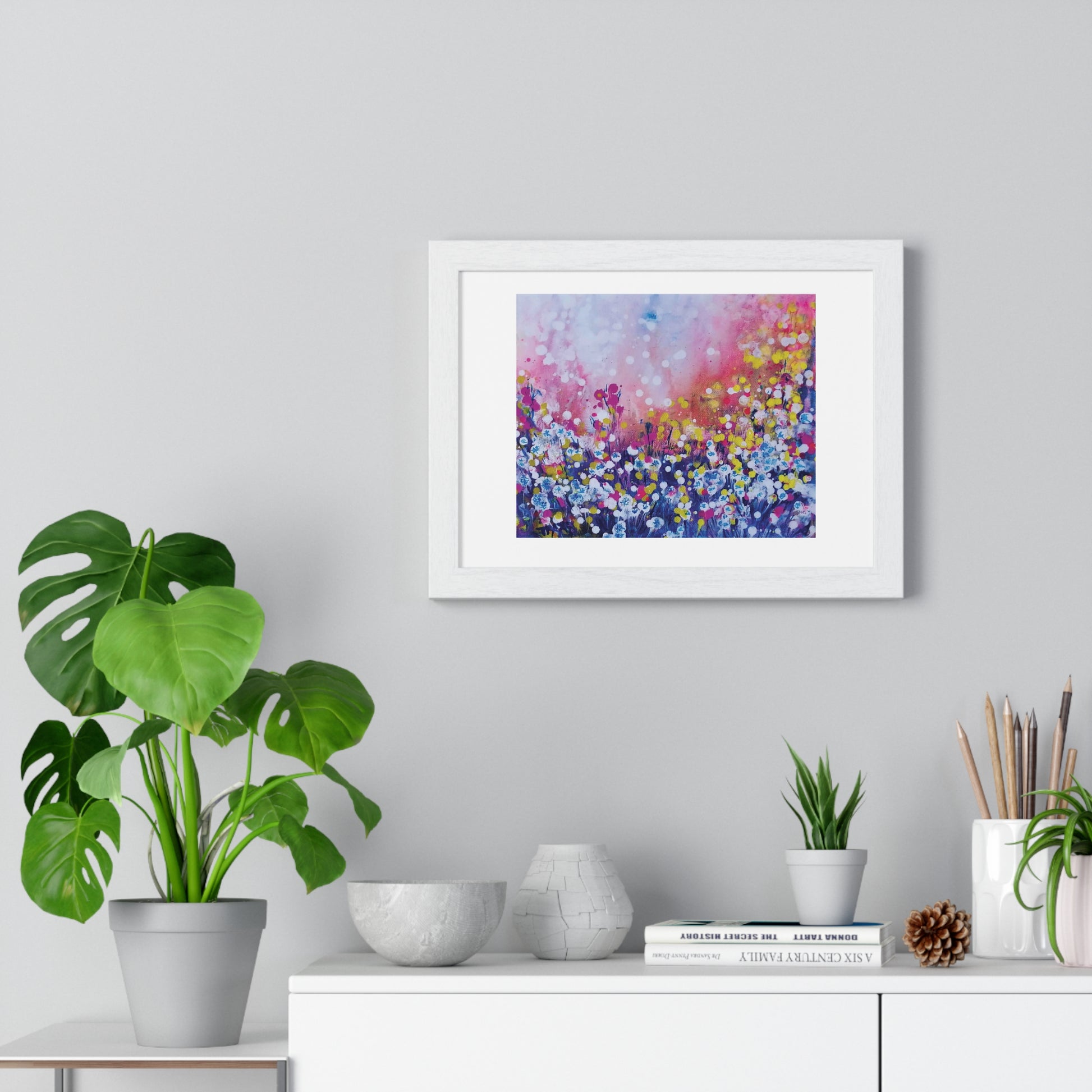 Abstract landscape in pink and blue with dandelions framed art print by contemporary artist TRAXI, featuring a magical sunrise scene on archival matte paper. A framed abstract floral painting with pink, purple, blue, and yellow tones hanging on a light gray wall above a white console table with plants and decor.