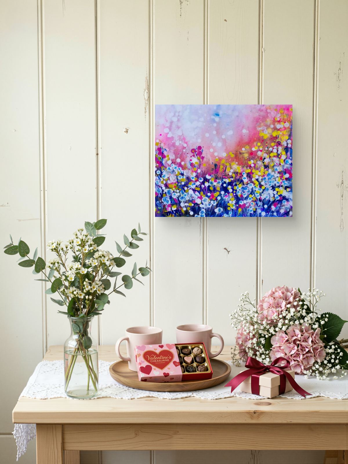 Original Painting, Life is Wonderful, Abstract Landscape, Dandelions Art, Pink and Blue Abstract, Small Art Gift, Diana Dimova TRAXI, Romantic Painting, Contemporary Fine Art, 30x25 cm A colorful floral canvas painting hangs on a cream-colored wooden paneled wall above a light wood table styled with pink mugs, chocolates, baby's breath, and pink hydrangeas.
