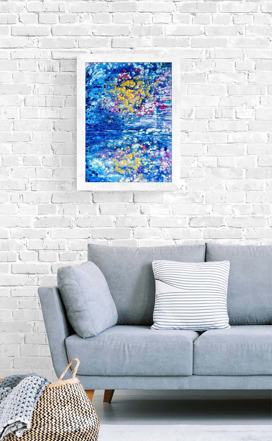 Abstract Reflections Sunset Seascape limited edition print in blue, pink, and gold by contemporary artist Diana Dimova TRAXI. Abstract art print featuring vibrant blue, yellow, and purple brushstrokes, displayed on a white brick wall above a light gray sofa with decorative pillows.