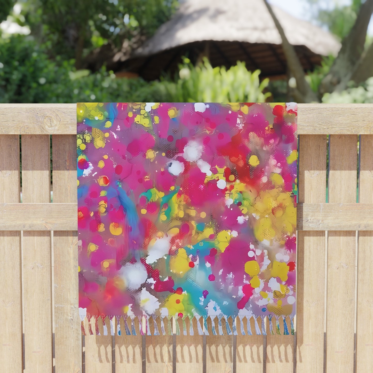 Boho beach towel of happiness featuring abstract wearable art inspired by the Paintings of Happiness series by Diana Dimova TRAXI. A square scarf with colorful paint splatters and a fringed bottom edge, draped over a light wood fence in an outdoor garden setting.