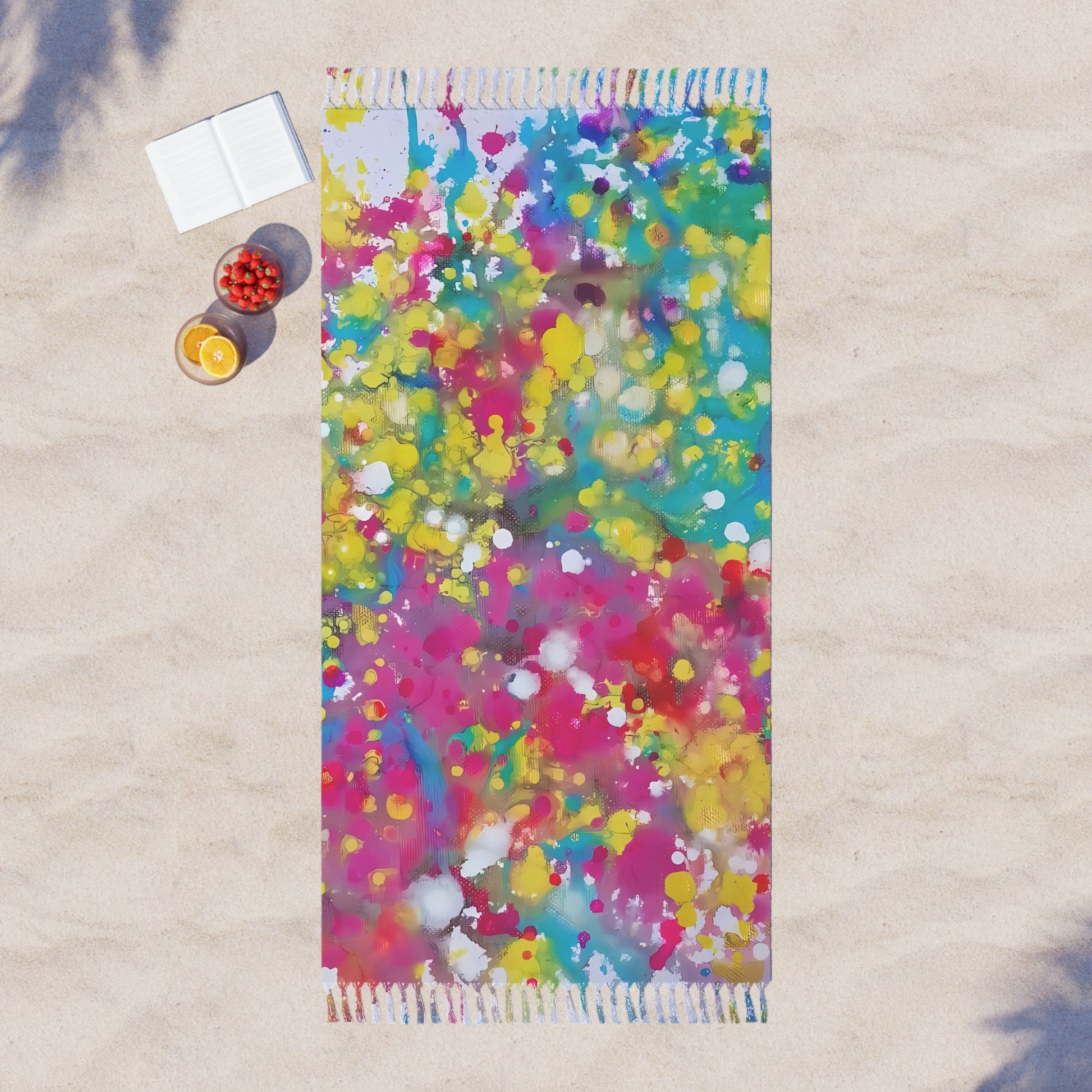 Boho beach towel of happiness featuring abstract wearable art inspired by the Paintings of Happiness series by Diana Dimova TRAXI. A rectangular beach towel with colorful paint splatter patterns lies on light beige sand next to a small white notebook and two small bowls of fruit.