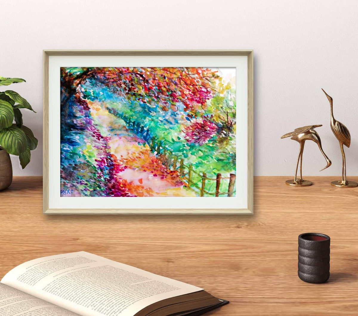 Colorful Autumn Landscape, original fall watercolor painting with an autumn path on paper by contemporary artist TRAXI. A framed colorful landscape painting of a tree-lined path, displayed on a wooden desk with a book, a potted plant, and two gold bird figurines against a light wall.