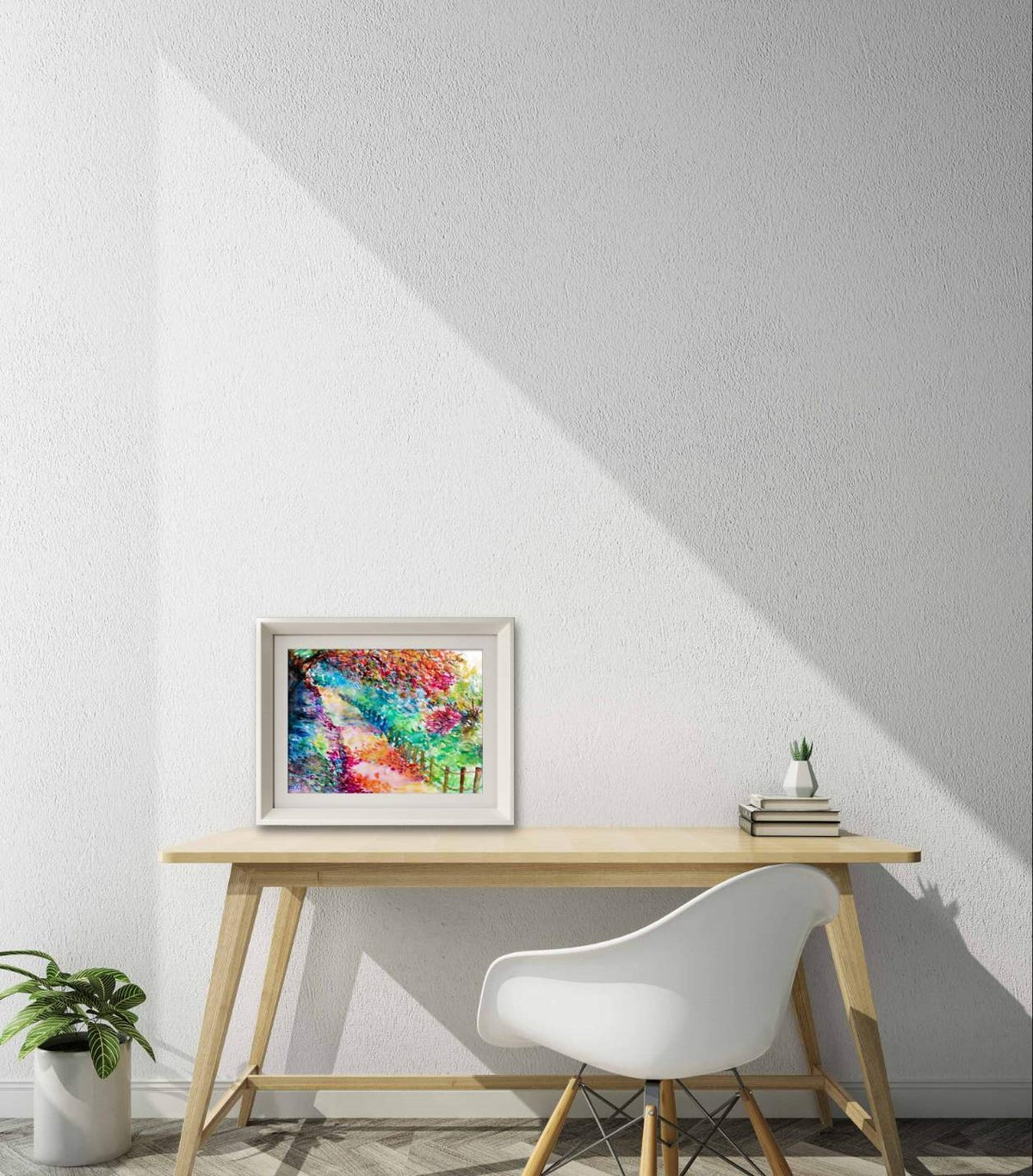 Colorful Autumn Landscape, original fall watercolor painting with an autumn path on paper by contemporary artist TRAXI. A colorful abstract framed art print is displayed on a light wood desk in a modern room setting with a white chair, a small potted plant, and books, under a strong diagonal shadow on a textured white wall.
