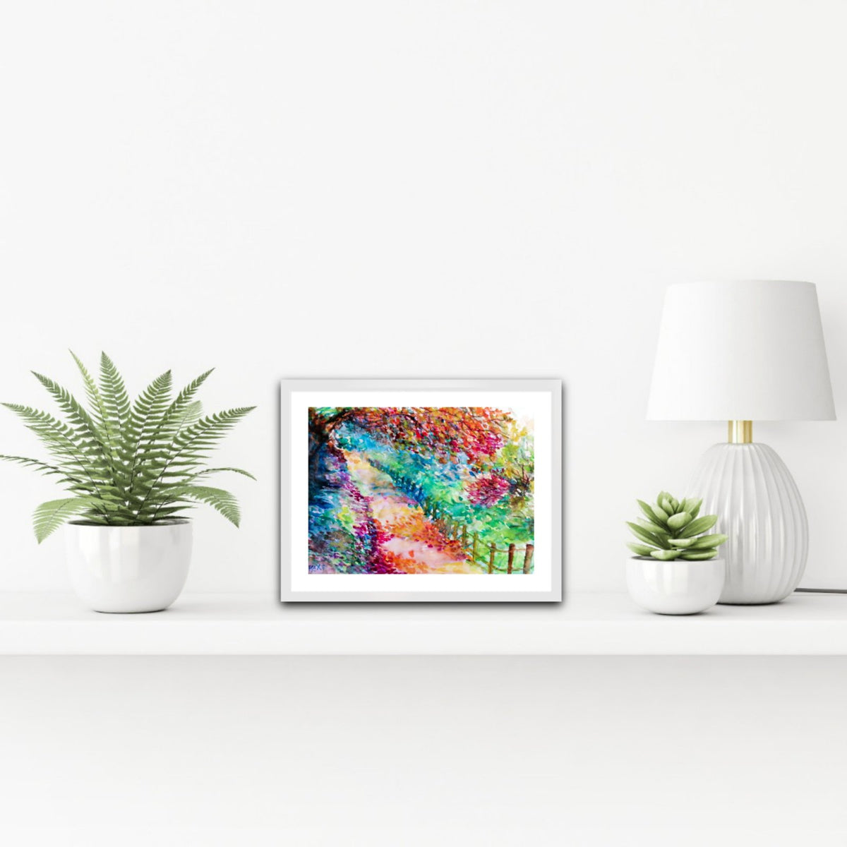 Colorful Autumn Landscape, original fall watercolor painting with an autumn path on paper by contemporary artist TRAXI. A framed abstract colorful painting sits on a white shelf between a potted fern, a small succulent, and a white table lamp against a white wall.