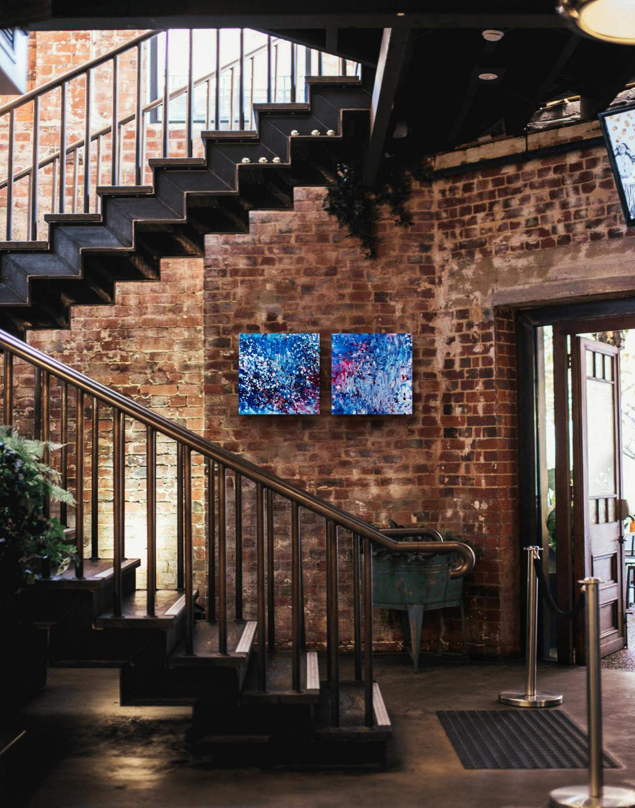 Two blue abstract energy canvas paintings by TRAXI ART curated as a diptych on a rustic brick wall in a modern commercial restaurant or hospitality interior design project.