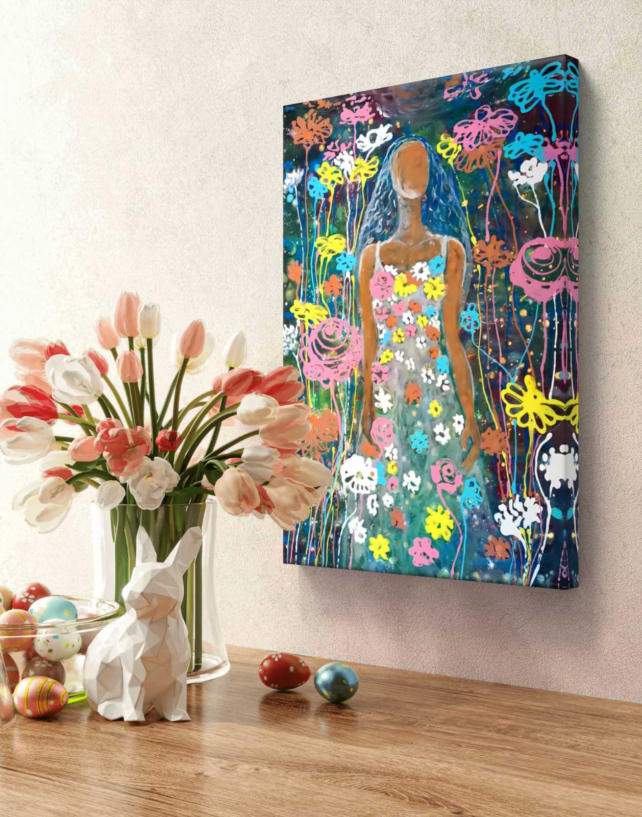 A colorful canvas wall art print depicting a stylized woman surrounded by vibrant flowers, displayed on a beige wall above a wooden table with a vase of tulips and decorative Easter eggs. Palm Sunday original spring abstract fluid acrylic painting with vibrant floral colors celebrating feminine energy and Bulgarian heritage by Diana Dimova TRAXI ART.