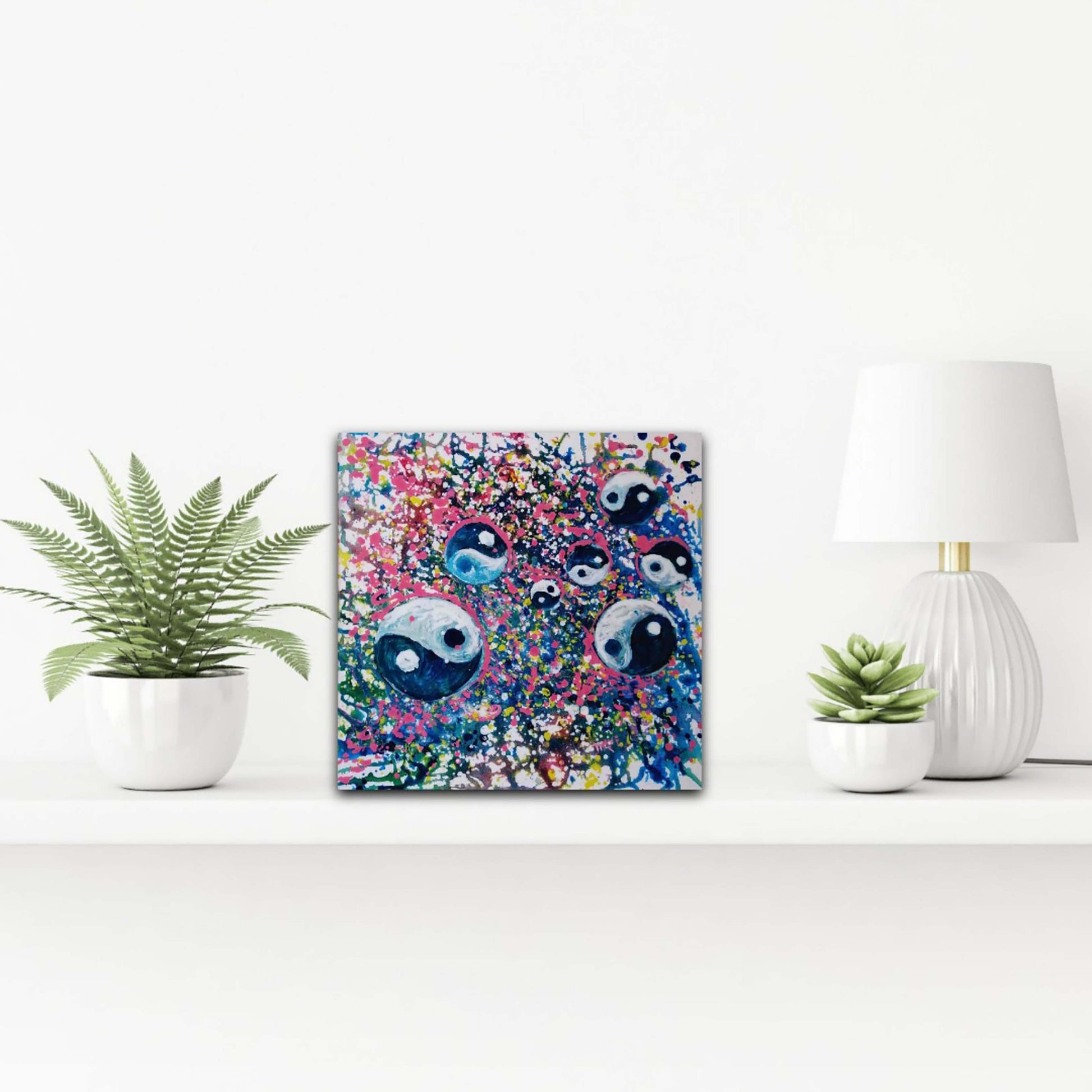 Yin Yang spiritual abstract painting titled Events of Life by Diana Dimova TRAXI featuring a circular blue, white, and soft pink composition for modern luxury interiors. A square canvas art print featuring a colorful, abstract design of cartoon eyes, styled on a white shelf next to potted plants and a white lamp.