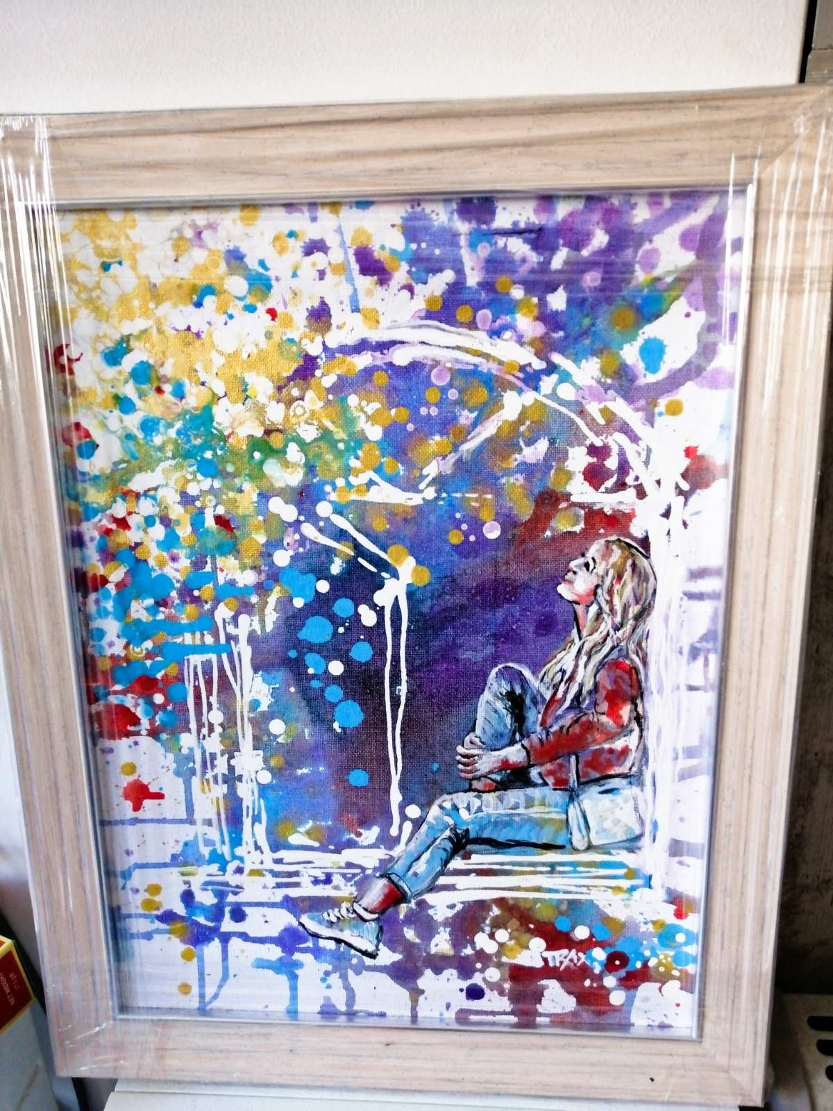 Original framed painting Talking with the Stars by Diana Dimova TRAXI, featuring a spiritual window composition with a girl reaching toward a starry night sky. A framed abstract watercolor painting featuring a seated woman in a red top and blue jeans under a colorful arch of splattered paint in purple, blue, yellow, red, and white. The artwork is housed in a light natural wood frame with a clear protective plastic cover.
