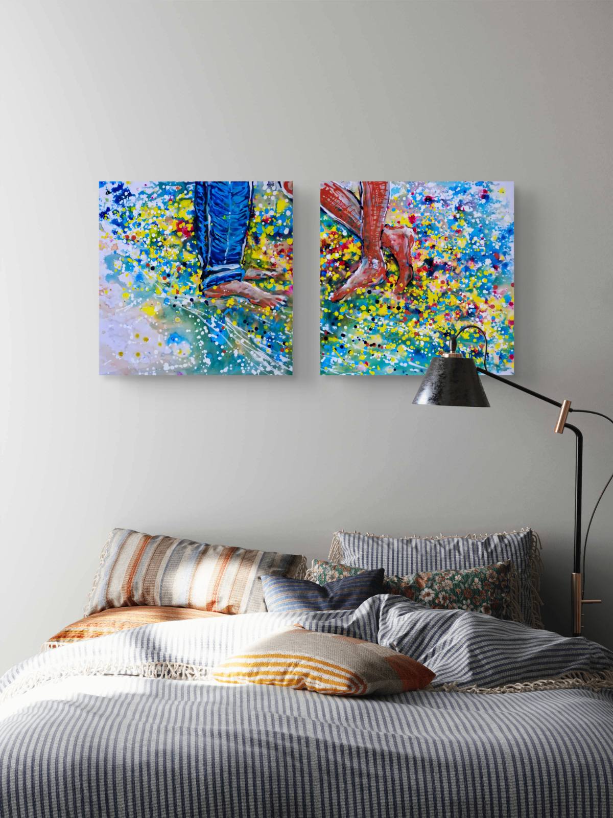 Love affair diptych original abstract painting set by contemporary artist TRAXI, depicting playful feet on the beach with vibrant fluid acrylic colors. A bedroom scene with a bed made up in striped bedding and pillows, a black floor lamp, and two colorful abstract wall art canvases above the bed. The canvases depict legs and boots in a vibrant blue, yellow, and red paint-splatter style.