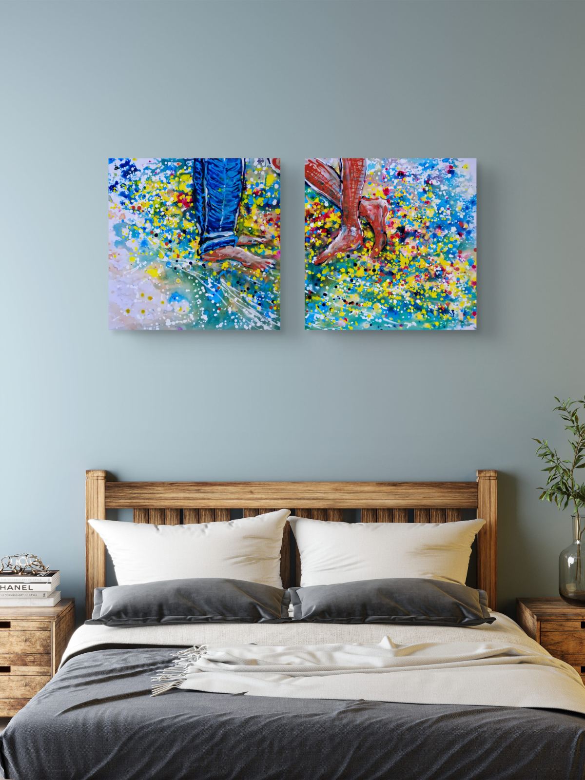 Love affair diptych original abstract painting set by contemporary artist TRAXI, depicting playful feet on the beach with vibrant fluid acrylic colors. A bedroom scene with a wooden bed dressed in white and gray bedding, and two colorful abstract canvas prints hanging on the wall above the headboard. The canvases depict legs walking through a vibrant field of blue, yellow, green, and red flowers.