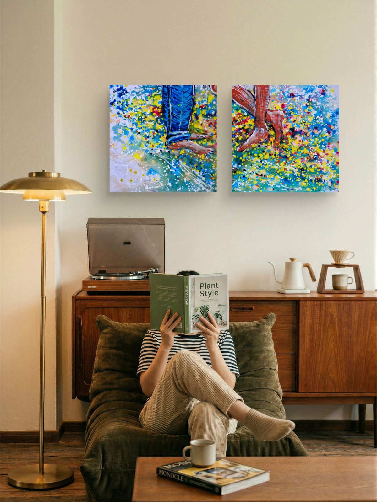 Love affair diptych original abstract painting set by contemporary artist TRAXI, depicting playful feet on the beach with vibrant fluid acrylic colors. A person sits in a green armchair reading a book in a cozy living room, with two colorful abstract canvas prints hanging on the wall above a wooden cabinet. A brass floor lamp stands to the left, and a coffee maker sits on the cabinet.