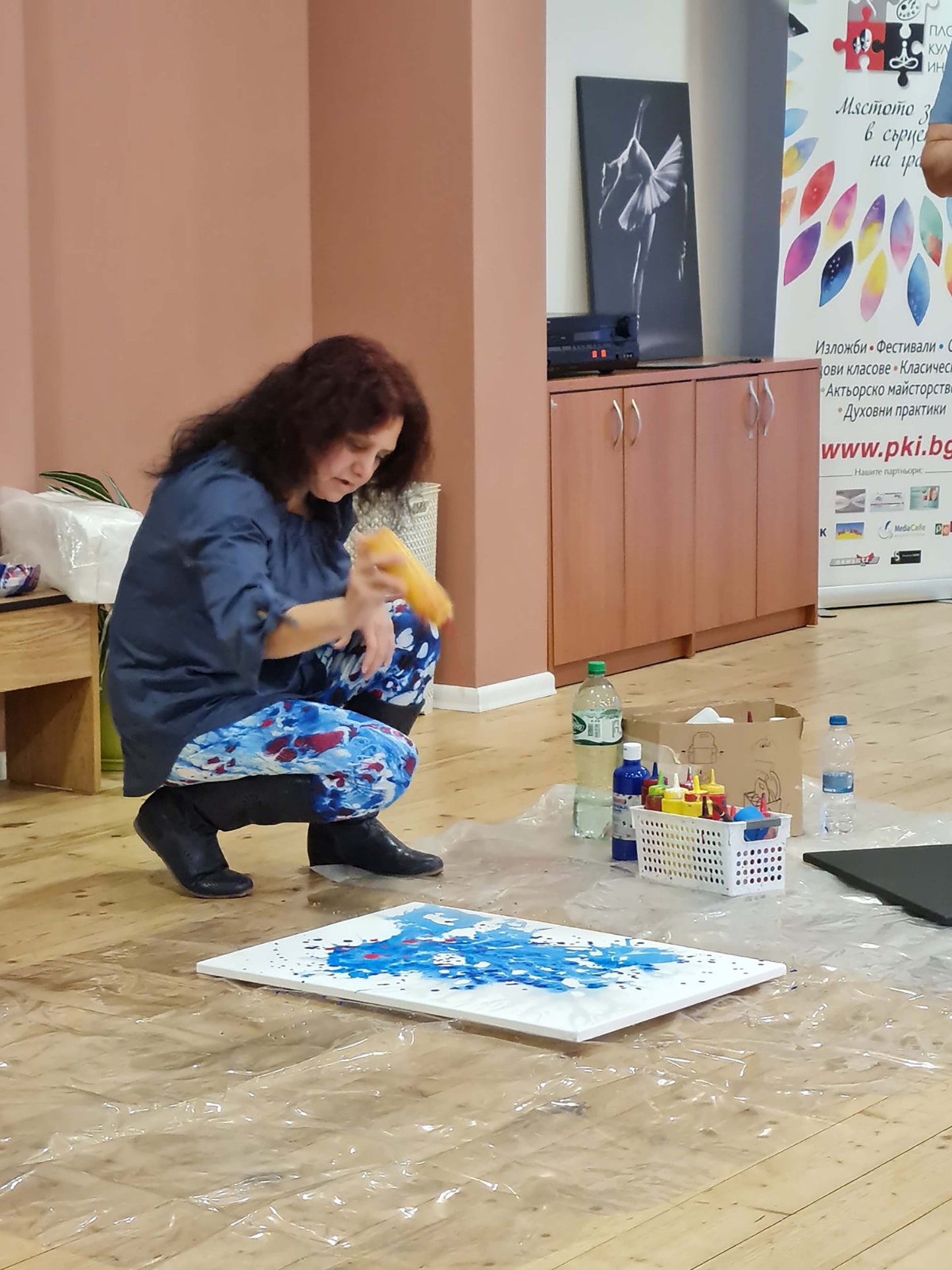 Mood of the Party original vertical abstract fluid acrylic painting with a vibrant blue wave and splashes of red and yellow by Diana Dimova TRAXI ART. A woman crouches on a wooden floor in an indoor studio or gallery space, painting on a white canvas with blue paint. She wears a dark blue shirt and patterned pants.