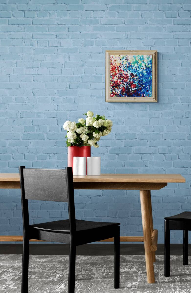 New Beginnings by contemporary artist TRAXI, an original abstract painting featuring blue, red, and gold fluid acrylic pouring techniques exploring emotional transformation. A styled interior scene with a light wood dining table, black chairs, a red vase of white flowers, and a colorful abstract painting on a blue brick wall.