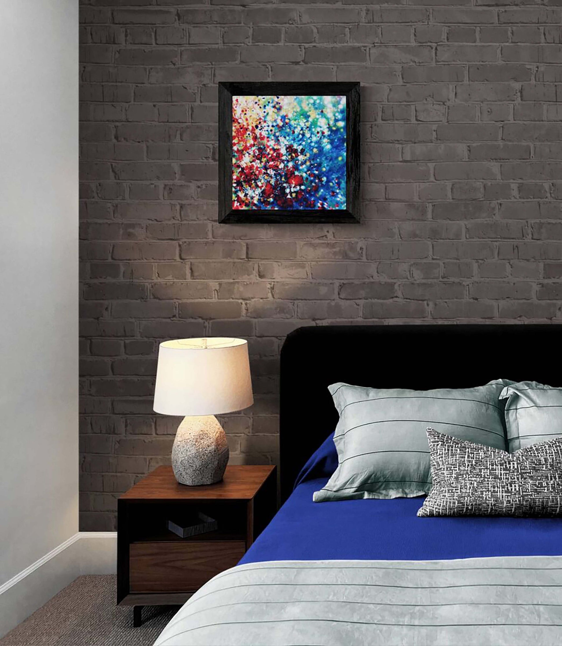 New Beginnings by contemporary artist TRAXI, an original abstract painting featuring blue, red, and gold fluid acrylic pouring techniques exploring emotional transformation. A framed abstract painting with vibrant red, blue, and white brushstrokes hangs on a dark gray brick wall above a bed with blue bedding and gray pillows in a modern bedroom setting.