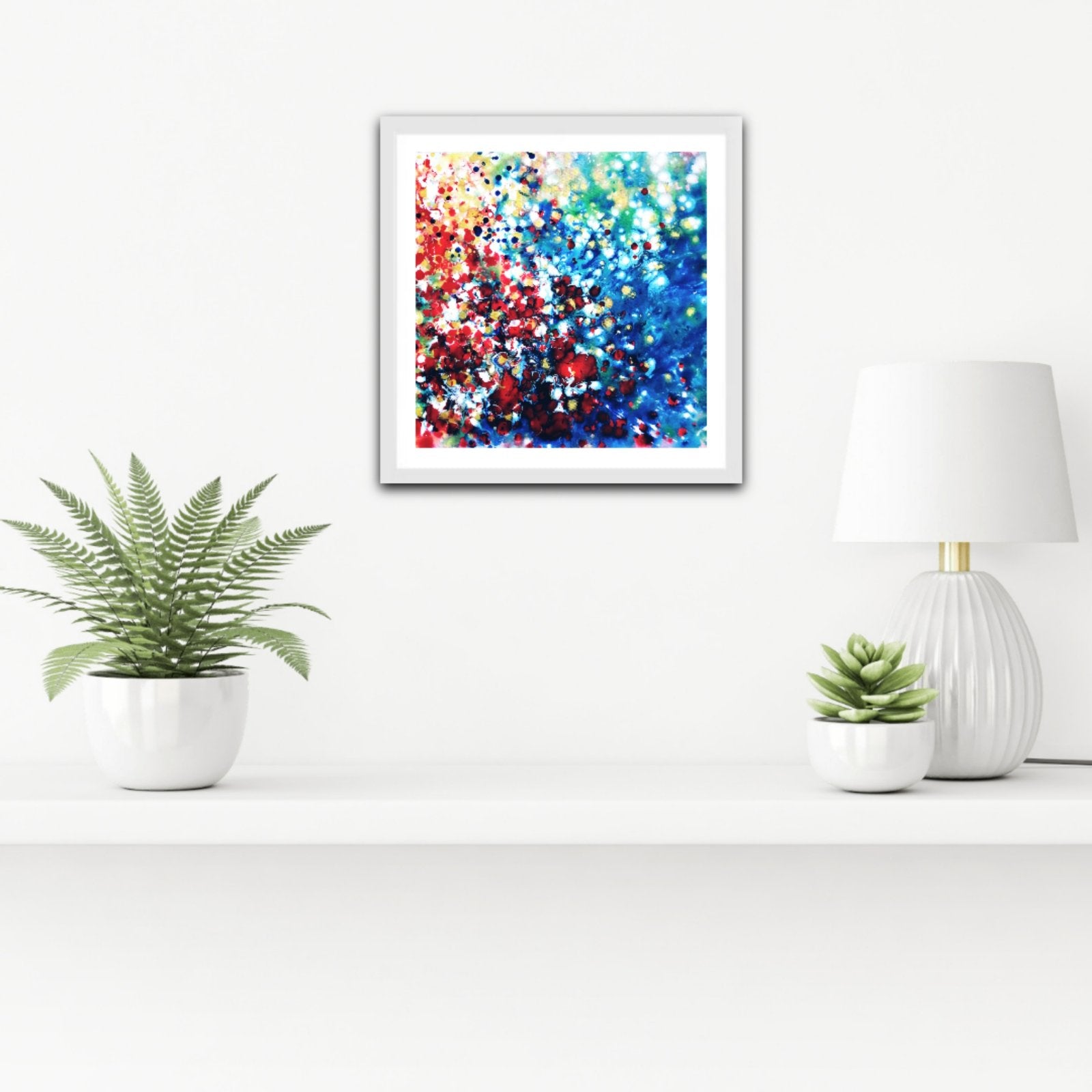 New Beginnings limited edition canvas print by contemporary artist TRAXI, featuring dynamic blue, red, and gold abstract acrylic pouring art exploring transformation. A framed abstract art print featuring a vibrant mix of blue, red, white, and gold bokeh-like dots, displayed on a white wall above a shelf with potted plants and a lamp.