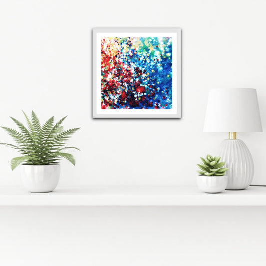 New Beginnings limited edition canvas print by contemporary artist TRAXI, featuring dynamic blue, red, and gold abstract acrylic pouring art exploring transformation. A framed abstract art print featuring a vibrant mix of blue, red, white, and gold bokeh-like dots, displayed on a white wall above a shelf with potted plants and a lamp.