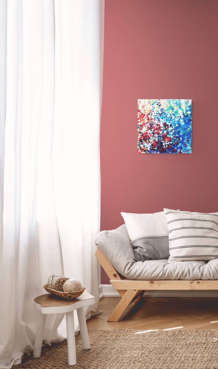 New Beginnings by contemporary artist TRAXI, an original abstract painting featuring blue, red, and gold fluid acrylic pouring techniques exploring emotional transformation. A colorful abstract canvas painting with blue, red, and white paint splatters, displayed on a dusty rose wall above a light wood daybed with gray and white pillows in a bright room.