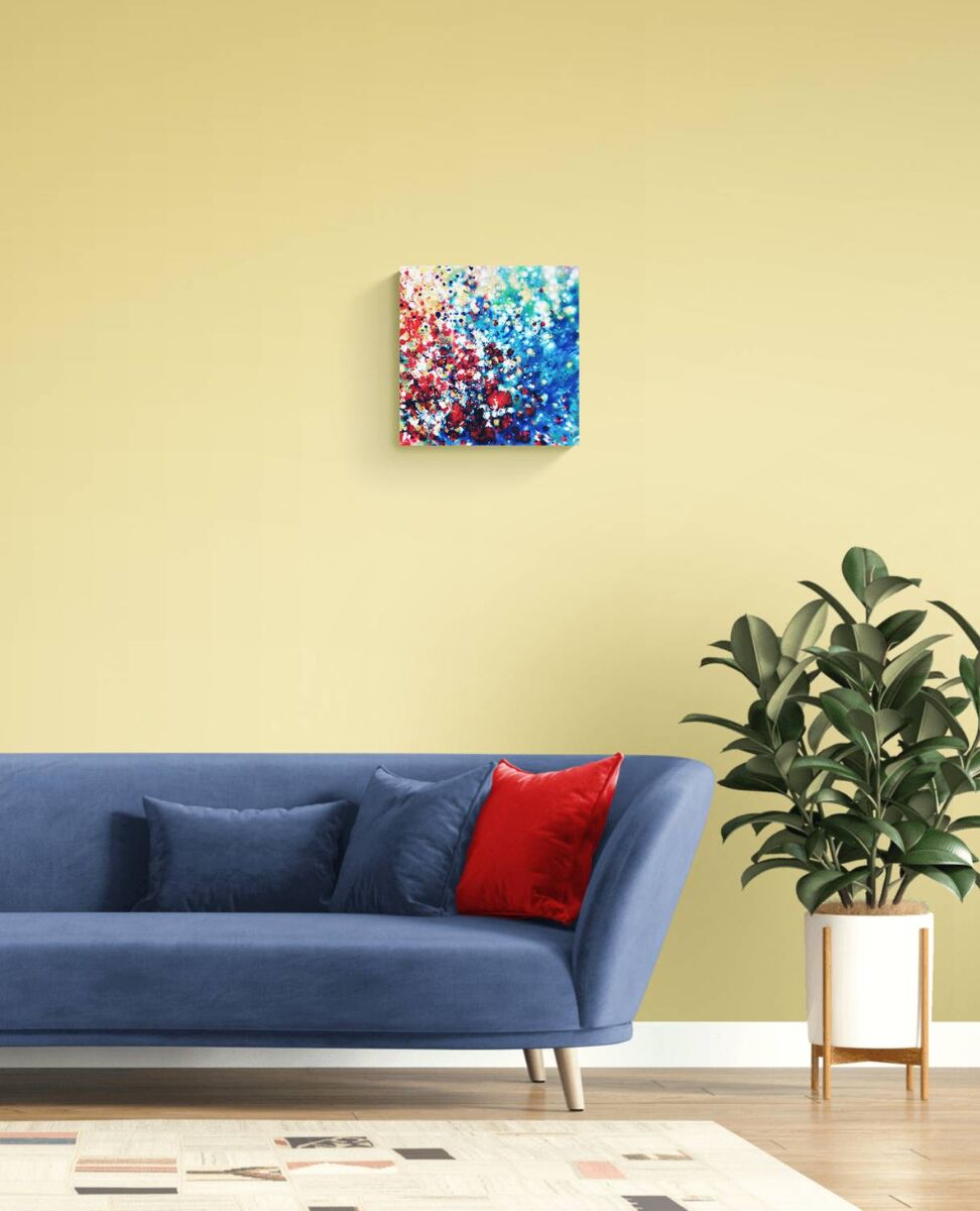 New Beginnings by contemporary artist TRAXI, an original abstract painting featuring blue, red, and gold fluid acrylic pouring techniques exploring emotional transformation. A square abstract canvas print with vibrant blue, red, and white paint splatters hanging on a pale yellow wall above a blue sofa with a red accent pillow, next to a potted green plant.
