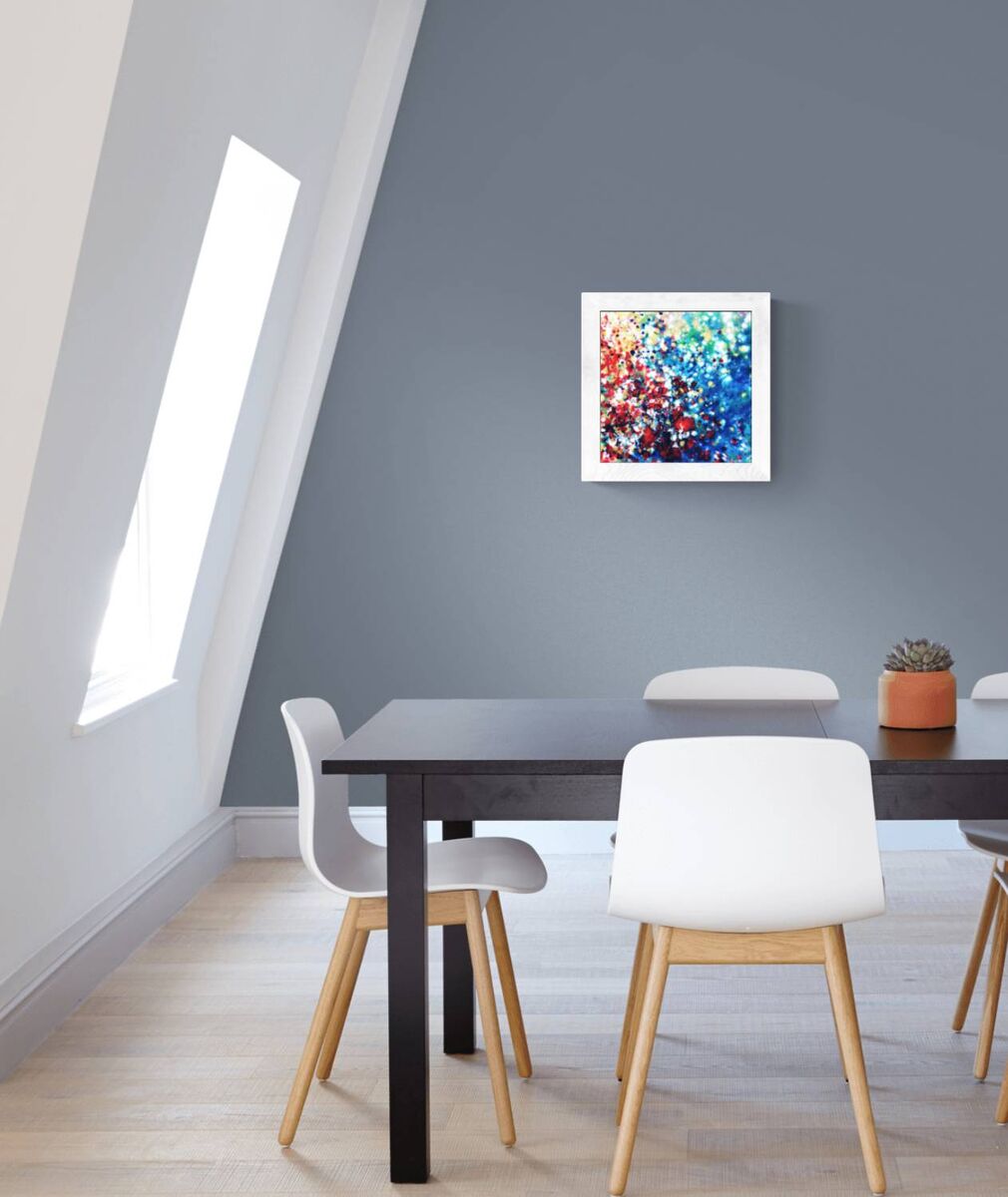 New Beginnings by contemporary artist TRAXI, an original abstract painting featuring blue, red, and gold fluid acrylic pouring techniques exploring emotional transformation. A square framed abstract painting with vibrant blue, red, and white brushstrokes hangs on a slate gray wall in a modern dining room setting with a black table and white chairs.