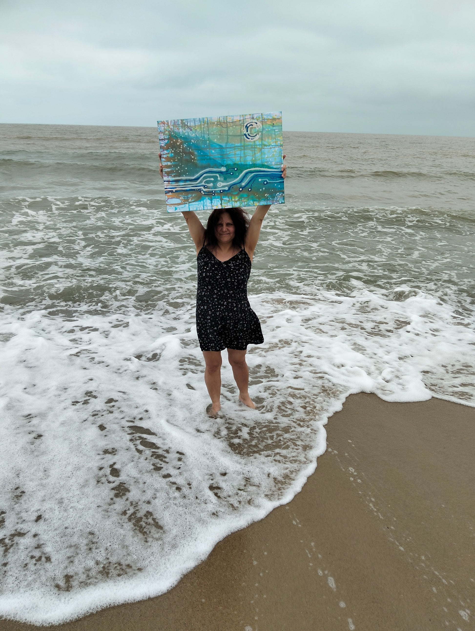 Seascape & Technology "Ocean Dialogue," an original contemporary abstract Blue  Ocean painting by Diana Dimova – TRAXI.  Artist TRAXI stands in the surf on a beach holding up a colorful square art print or painting with blue, green, and white abstract wave-like patterns.