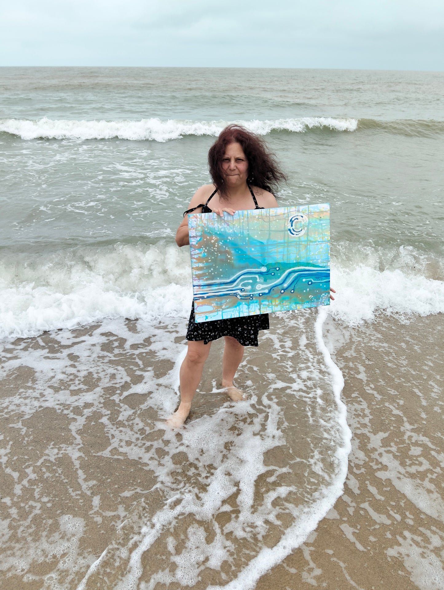 Seascape & Technology "Ocean Dialogue," an original contemporary abstract Blue  Ocean painting by Diana Dimova – TRAXI.  Artist TRAXI standing in shallow ocean waves on a beach holds up a square abstract painting featuring blue, teal, and white brushstrokes with a small circular logo in the upper right.
