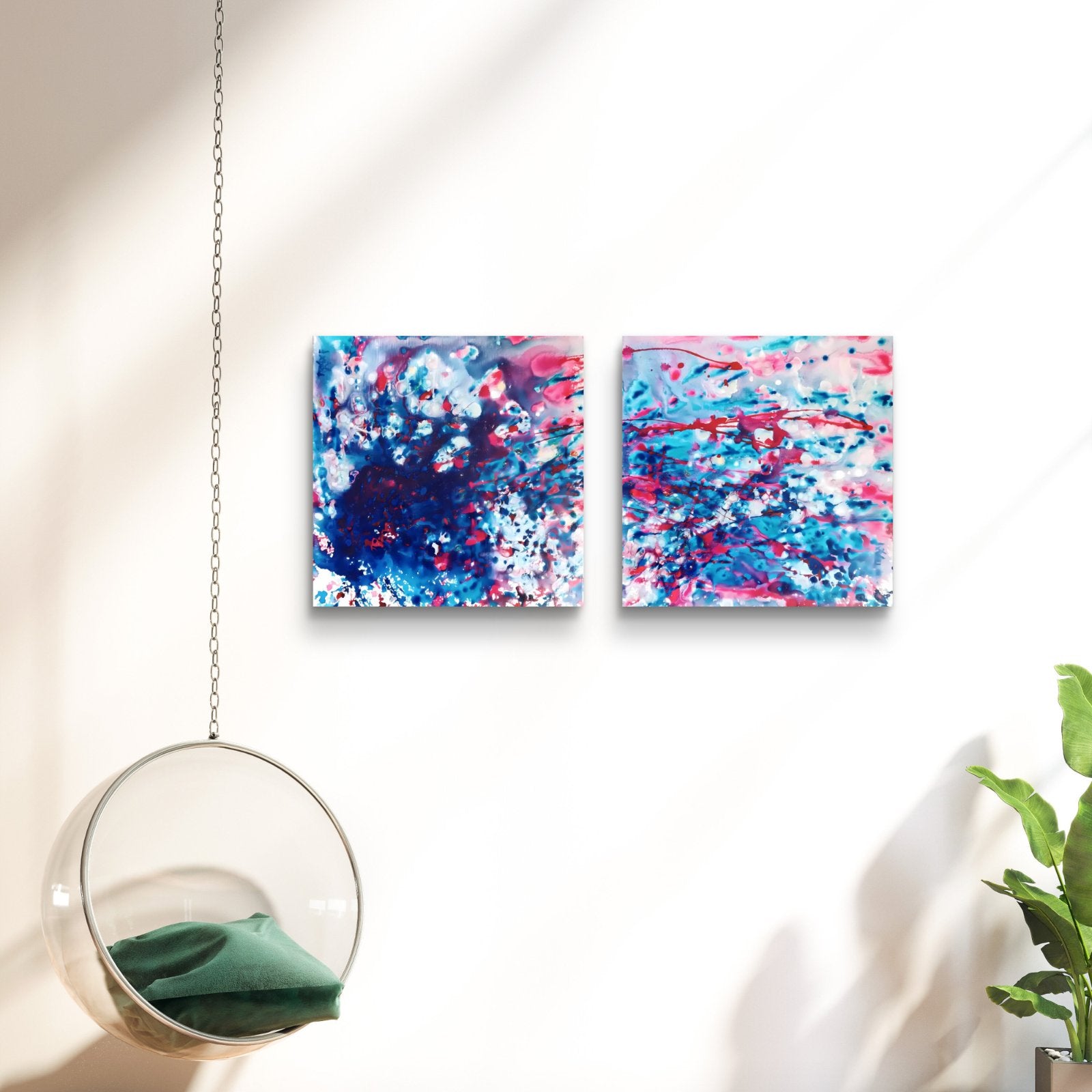 Original abstract diptych Theory of Attraction by Diana Dimova TRAXI, featuring blue, white, and rose tones exploring manifestation and cosmic synchronicity. Two square abstract wall art canvases with vibrant blue, pink, and white paint splatters, displayed side-by-side on a white wall in a modern room setting with a hanging glass chair and a green plant.