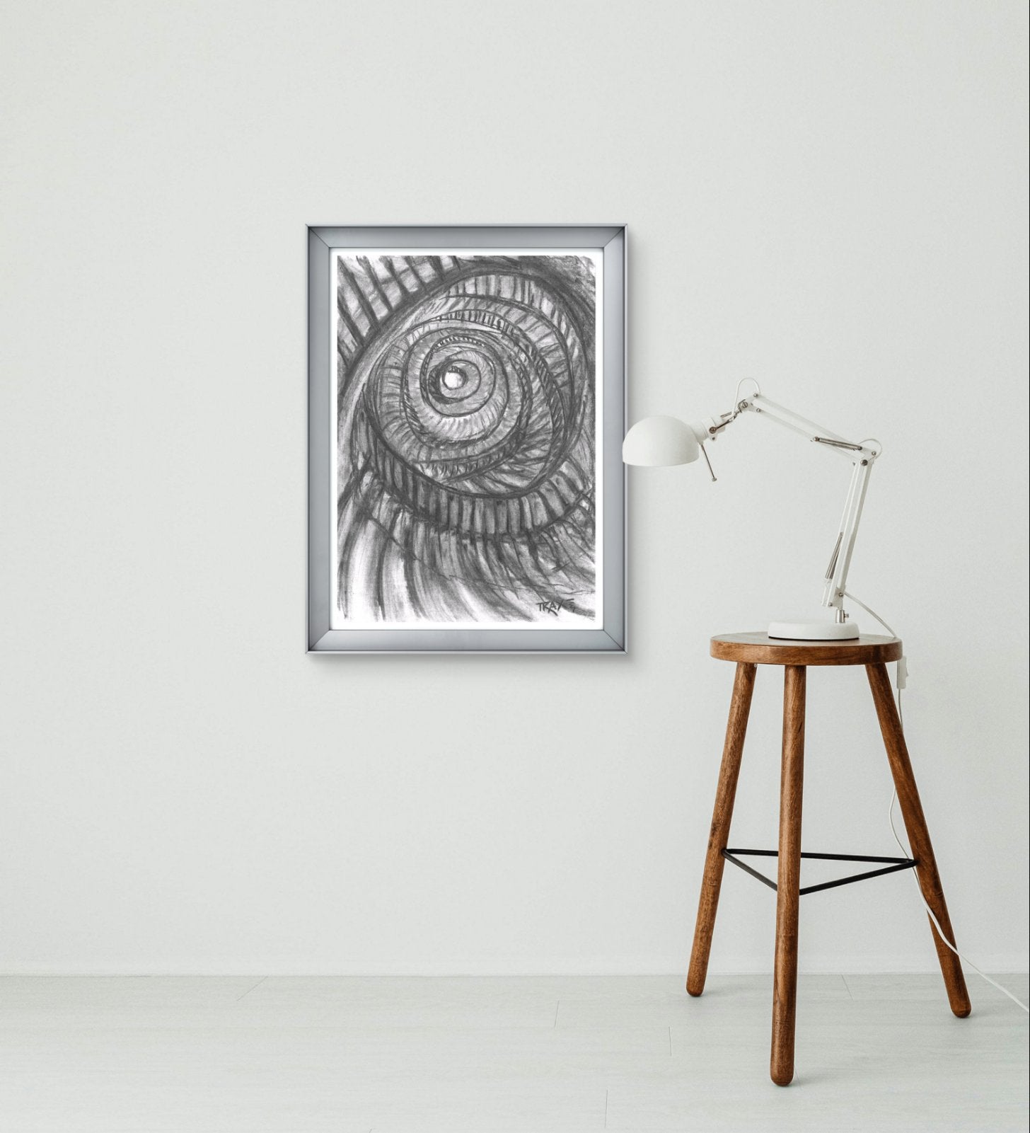 Original monochrome charcoal abstract drawing Eternal Spirals by Diana Dimova TRAXI, featuring rhythmic spiral movements in black and white tones. A framed black and white spiral art print hangs on a white wall in a minimalist room setting, next to a wooden stool with a white desk lamp.