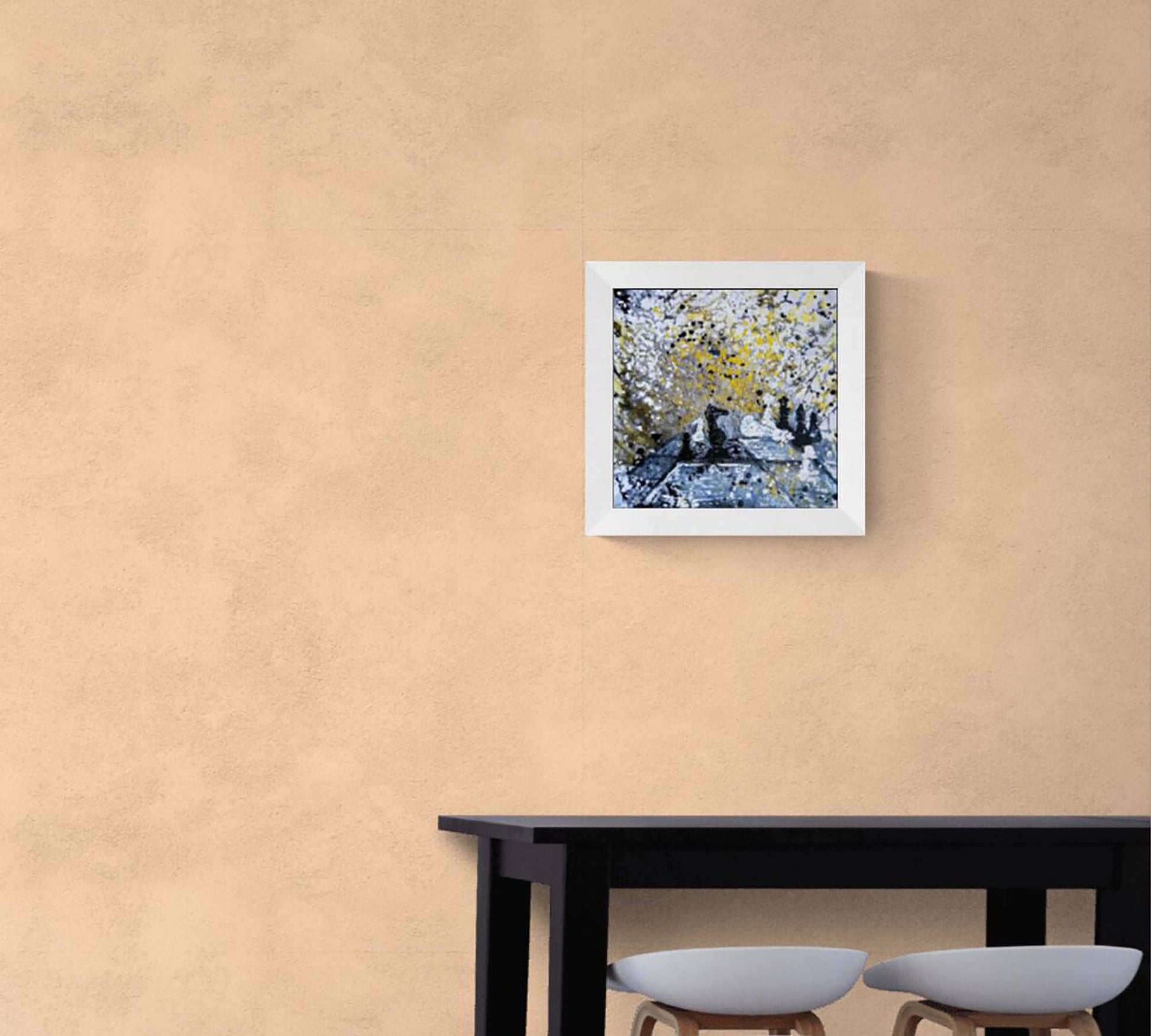 Original symbolic abstract painting Black Wins Again by Diana Dimova TRAXI, featuring chess elements in white, black, and gold tones. A square framed art print depicting a blue and yellow textured landscape hangs on a textured beige wall above a black table with two stools.