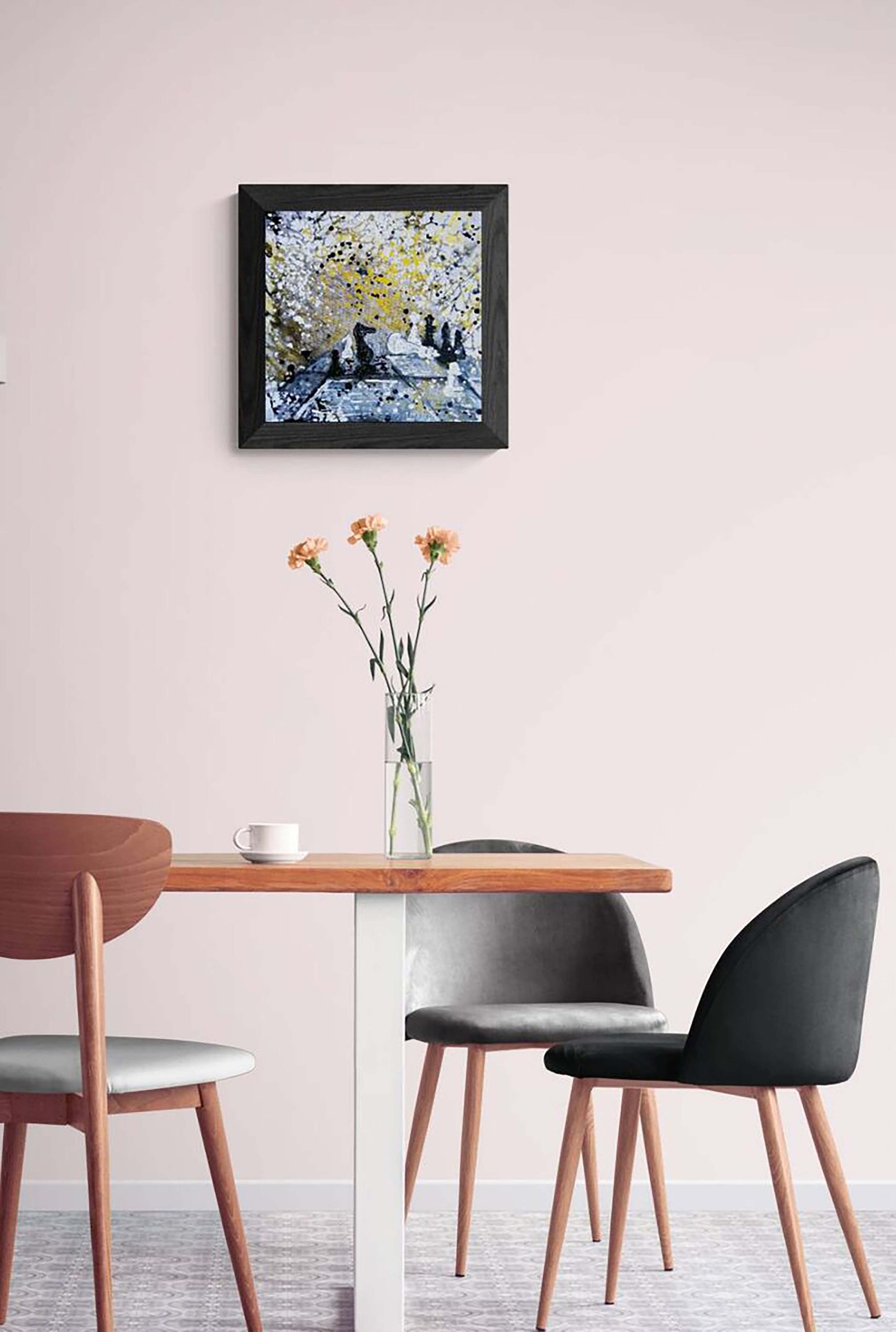 Original symbolic abstract painting Black Wins Again by Diana Dimova TRAXI, featuring chess elements in white, black, and gold tones. A framed abstract painting with blue, yellow, and white tones hangs on a pale pink wall above a wooden dining table with three chairs.