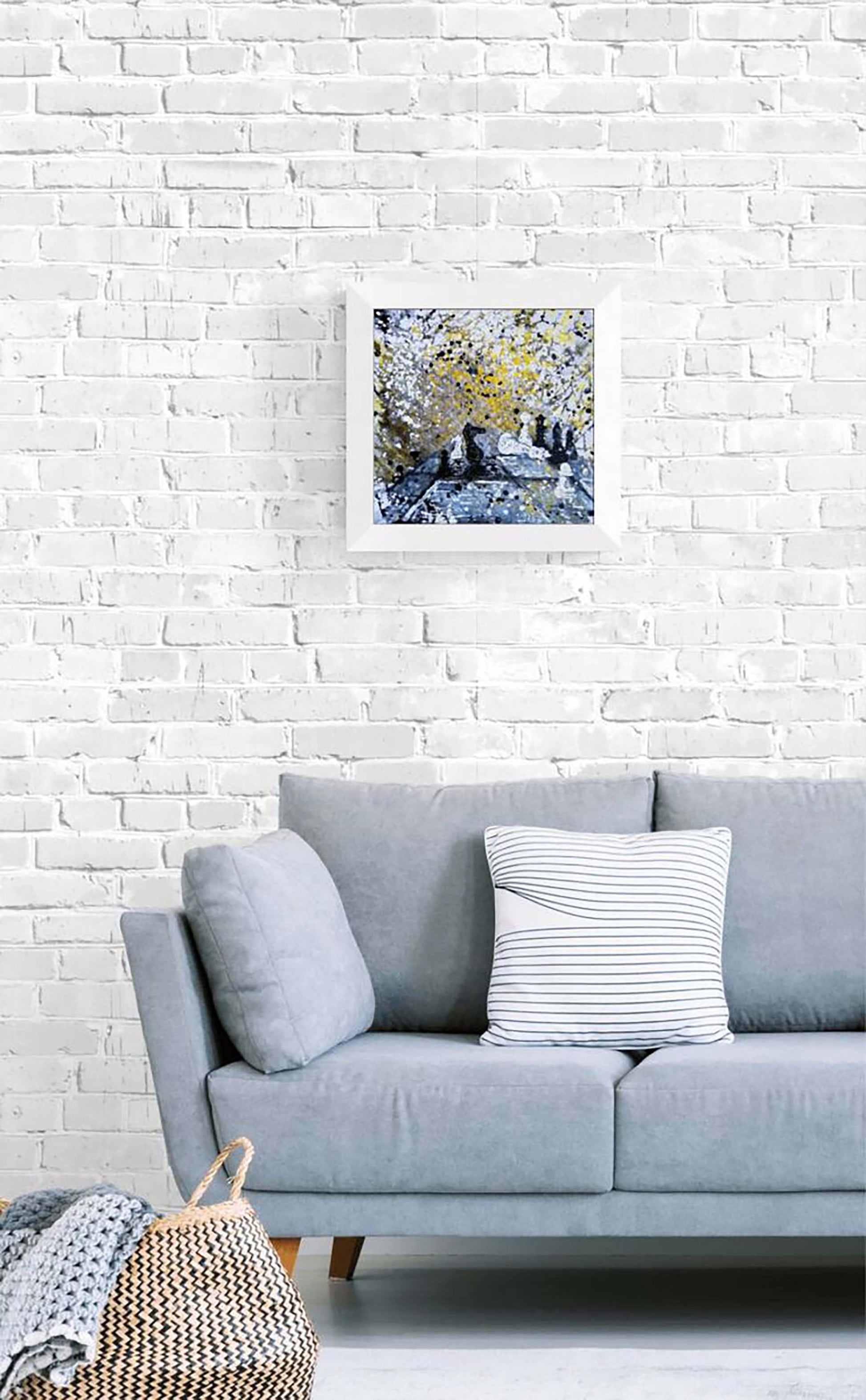 Original symbolic abstract painting Black Wins Again by Diana Dimova TRAXI, featuring chess elements in white, black, and gold tones. A framed abstract landscape art print with blue, yellow, and white tones hangs on a white brick wall above a light gray sofa with decorative pillows.