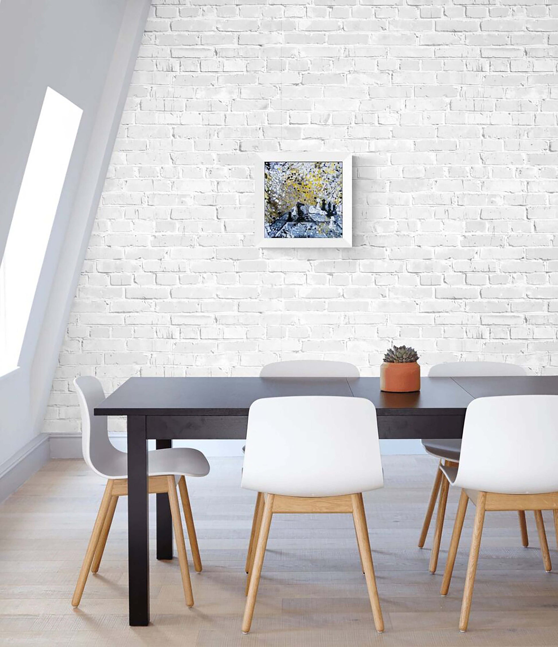 Original symbolic abstract painting Black Wins Again by Diana Dimova TRAXI, featuring chess elements in white, black, and gold tones. A framed art print depicting a scenic landscape with trees and water is displayed on a white brick wall above a dining table with white chairs in a bright, modern room.