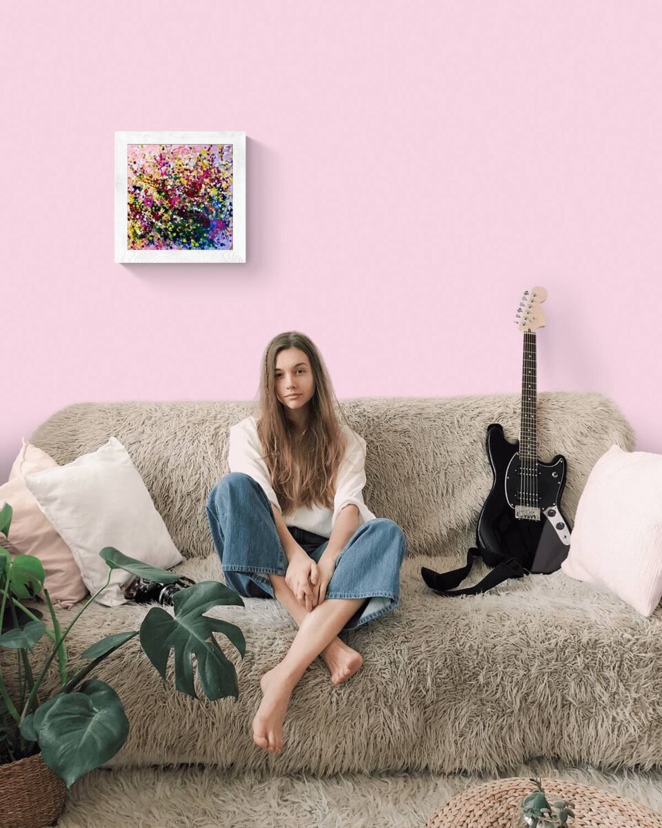 Original abstract painting Summer Garden Flowers by Diana Dimova TRAXI, featuring a colorful floral bouquet in vibrant yellow, pink, and orange tones. A woman sits on a shaggy beige couch in a pink room, with a colorful abstract painting on the wall, a black electric guitar, and a potted plant in the foreground.