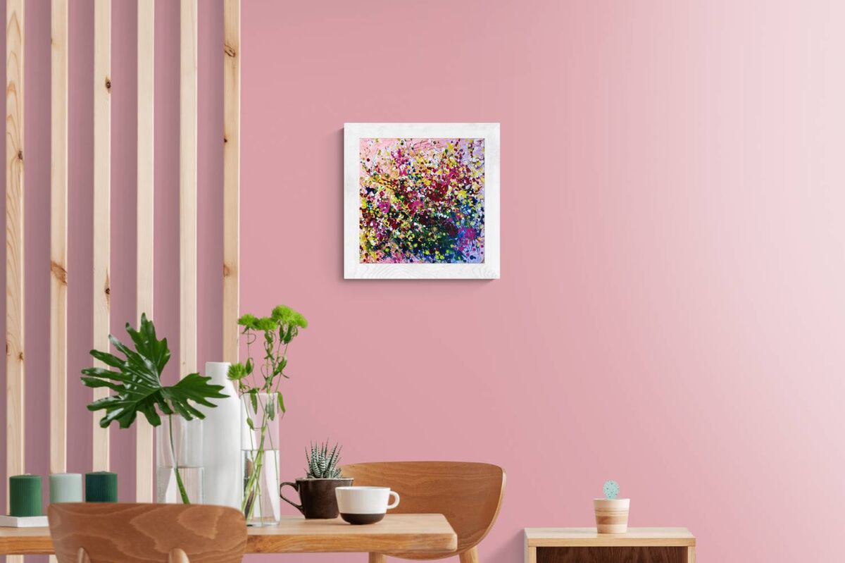 Original abstract painting Summer Garden Flowers by Diana Dimova TRAXI, featuring a colorful floral bouquet in vibrant yellow, pink, and orange tones. A colorful abstract floral art print in a white square frame, displayed on a pink wall in a modern dining room setting with wooden chairs, a table, and plants.