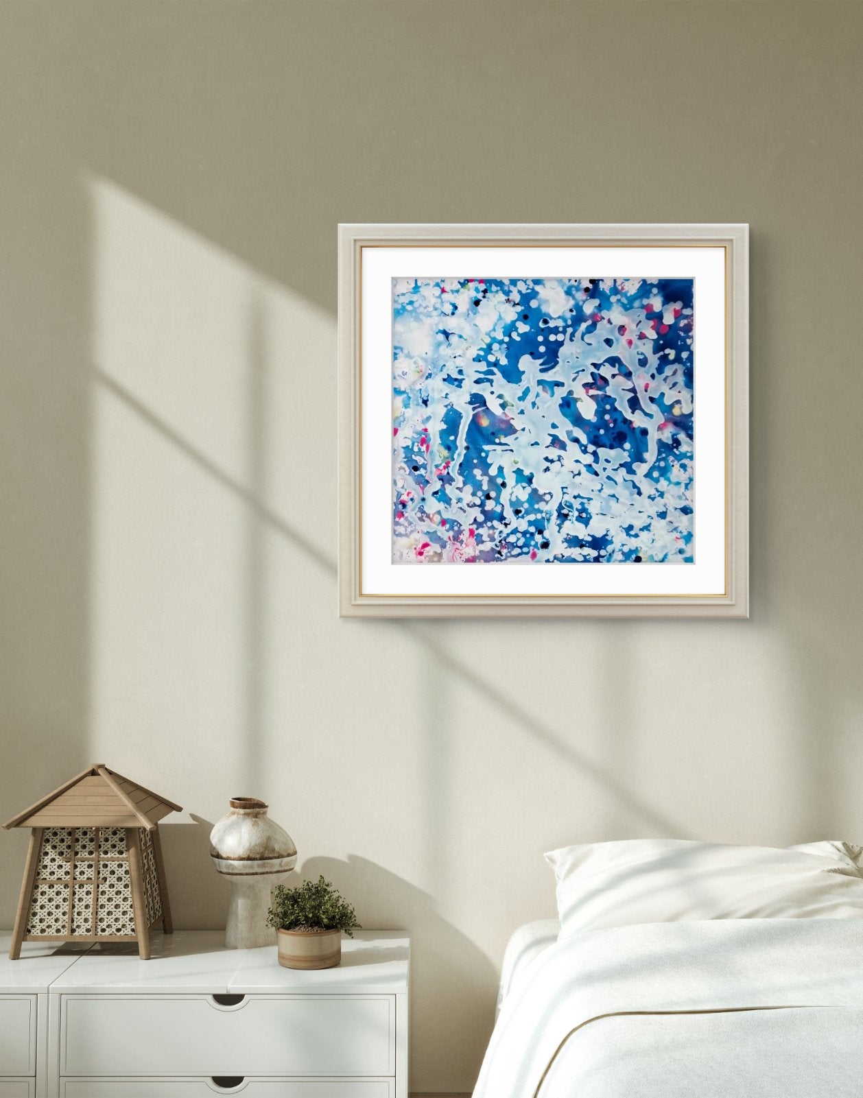 Original spiritual abstract painting Prayer by Diana Dimova TRAXI, featuring a white angel and protective wings in white and blue tones. Framed abstract wall art hanging above a bed in a softly styled bedroom. The artwork features a blue, white, and pink marbled or splattered pattern inside a light wood frame with a white mat.
