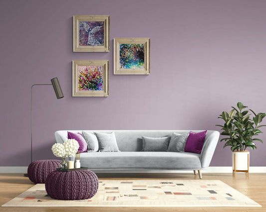 Original spiritual triptych Angelic Wall set by Diana Dimova TRAXI, featuring three connected fluid acrylic paintings representing protection and love. A styled living room scene with a light gray sofa, purple accent pillows, a patterned rug, a floor lamp, and a potted plant against a lavender wall with three framed abstract paintings.