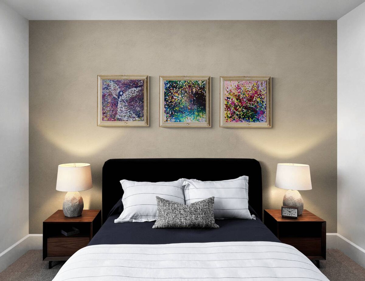 Original spiritual triptych Angelic Wall set by Diana Dimova TRAXI, featuring three connected fluid acrylic paintings representing protection and love. A styled bedroom interior featuring a bed with white and gray bedding, a black headboard, two matching wooden nightstands with lit table lamps, and three framed abstract paintings centered on a warm beige wall.