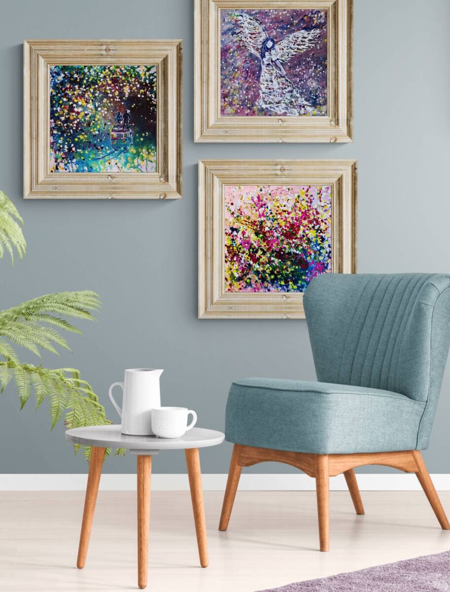 Original spiritual triptych Angelic Wall set by Diana Dimova TRAXI, featuring three connected fluid acrylic paintings representing protection and love. Three framed abstract paintings in light wood frames are displayed on a blue-gray wall above a teal armchair and a small round side table with a white pitcher and cup.